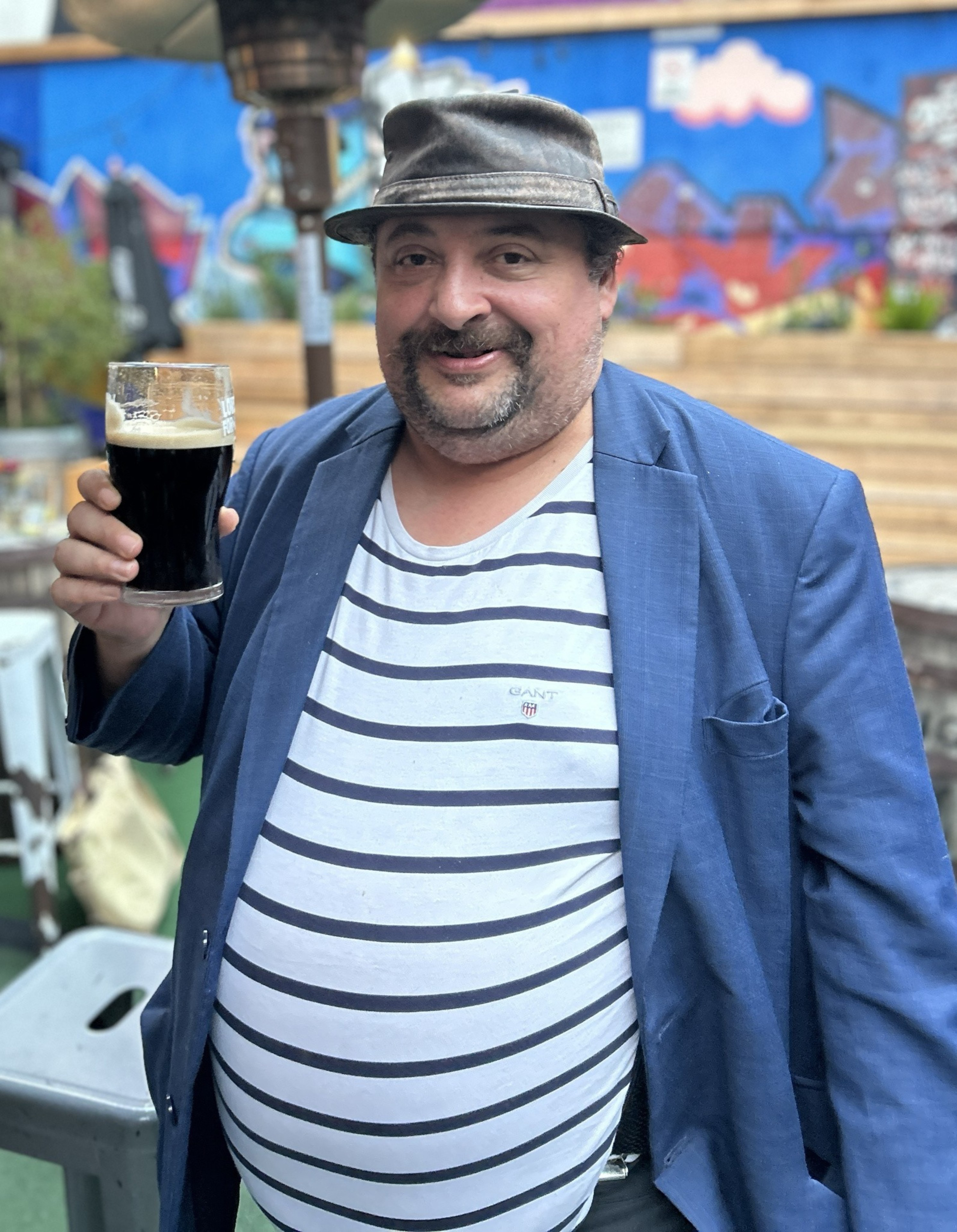 A man in a striped shirt and blue jacket holds a pint of dark beer. He's wearing a brown hat and standing in front of a colorful, graffiti-covered wall.