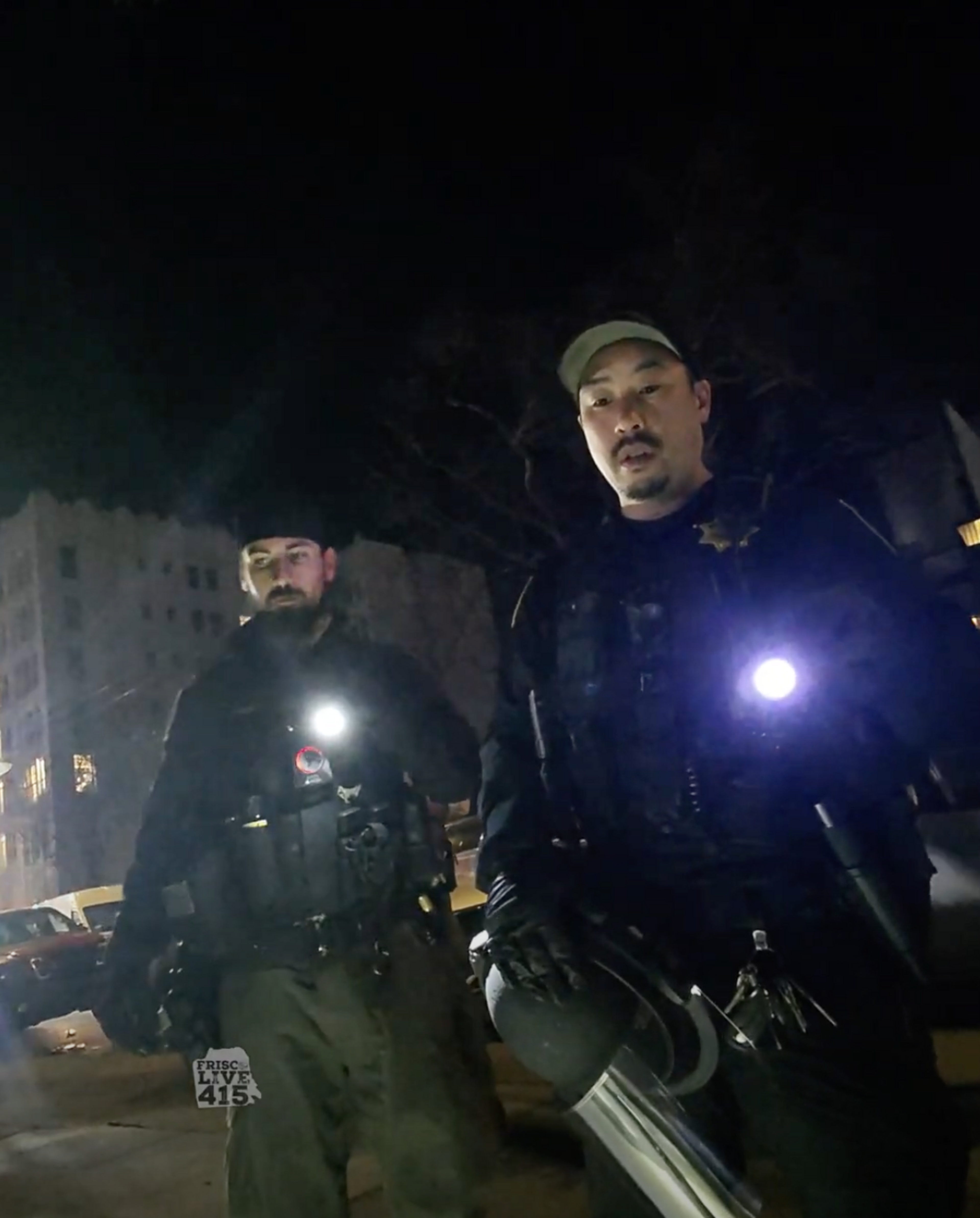 Two officers stand outdoors at night, holding flashlights. They wear tactical gear and appear to be on a street. A building is faintly visible in the background.