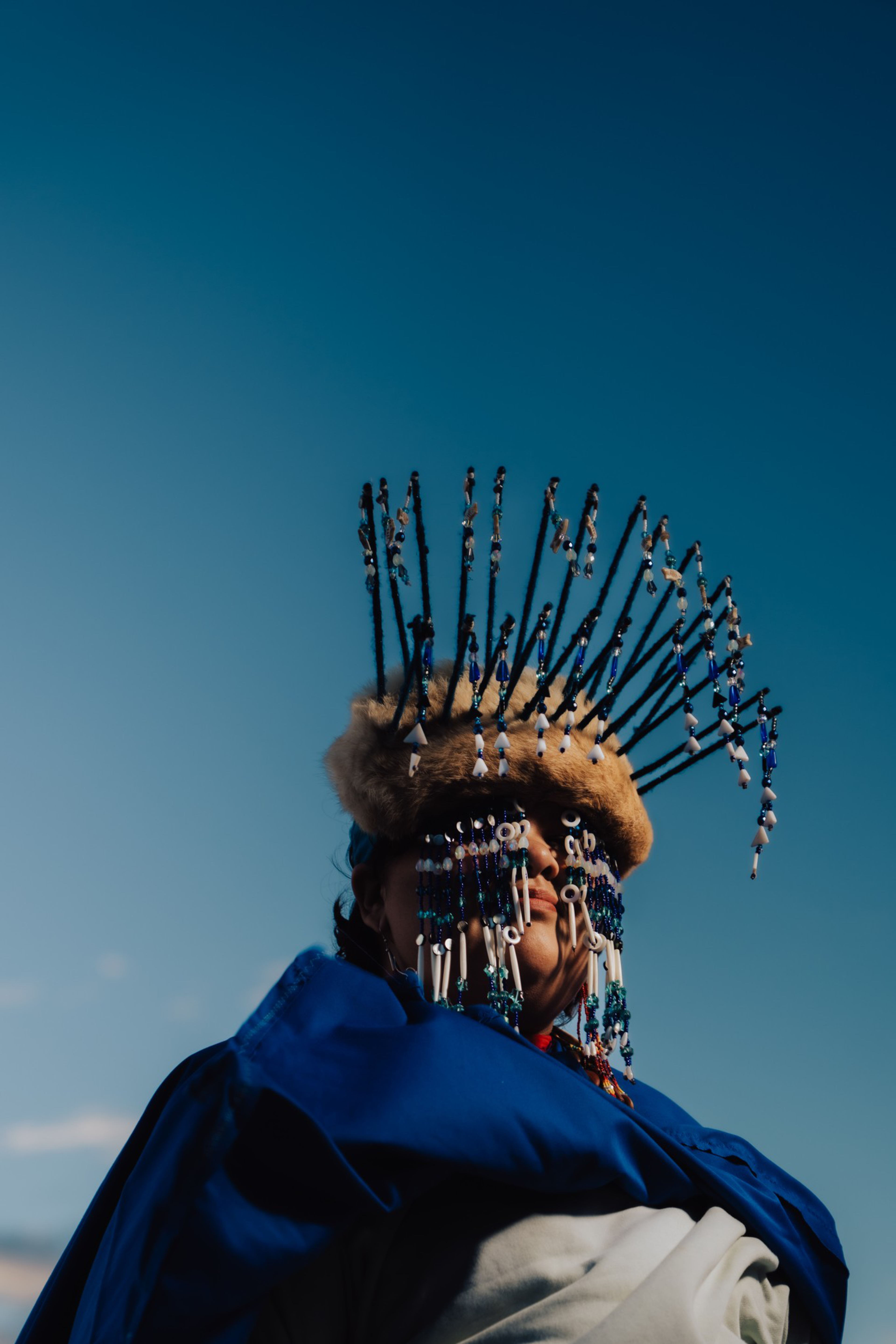 A person wears a fur hat adorned with tall beaded sticks and matching beaded face decorations, set against a clear blue sky.