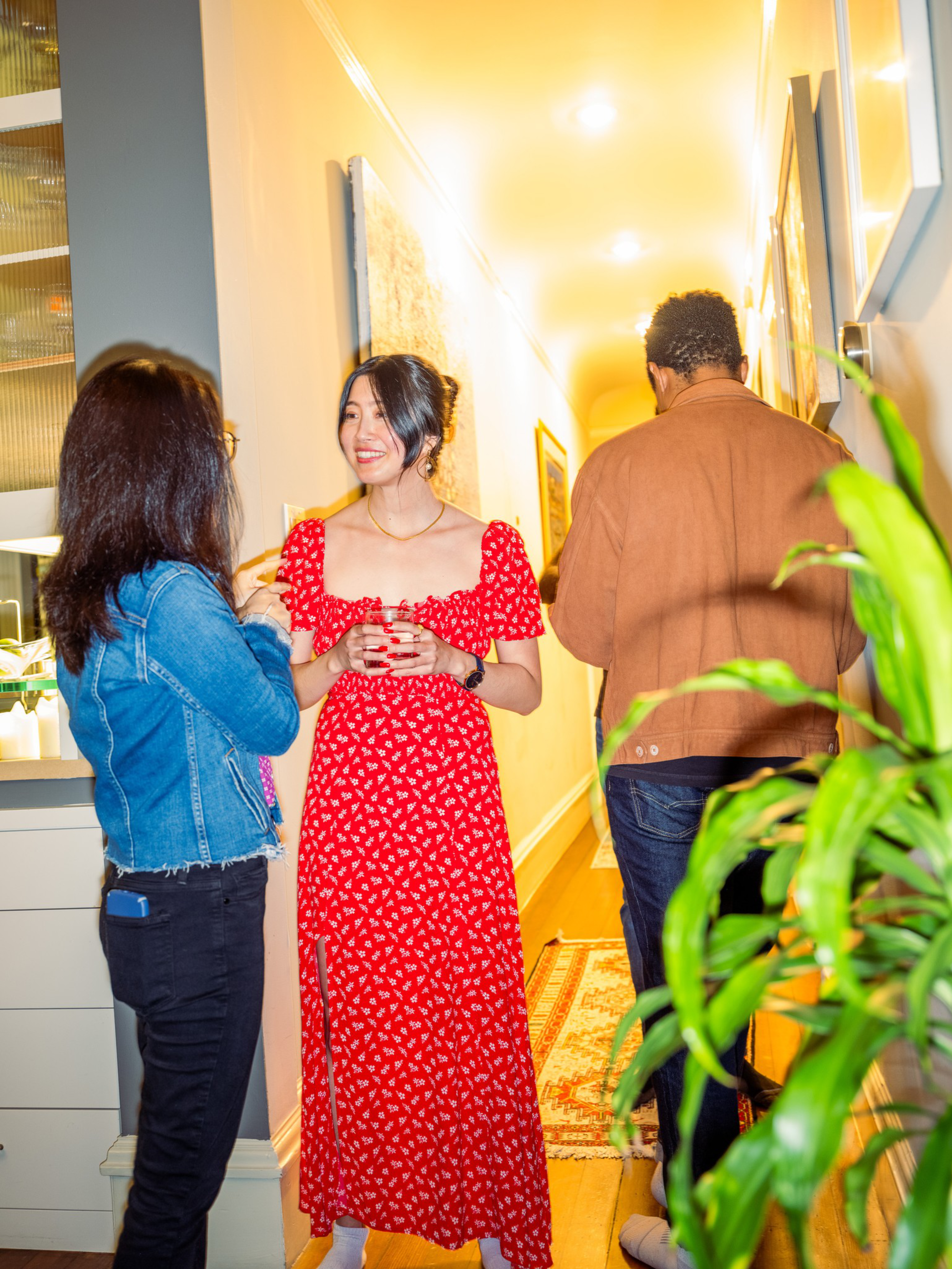 A woman in a red dress talks and smiles with a woman in a denim jacket, while a man in a brown jacket walks down a hallway.