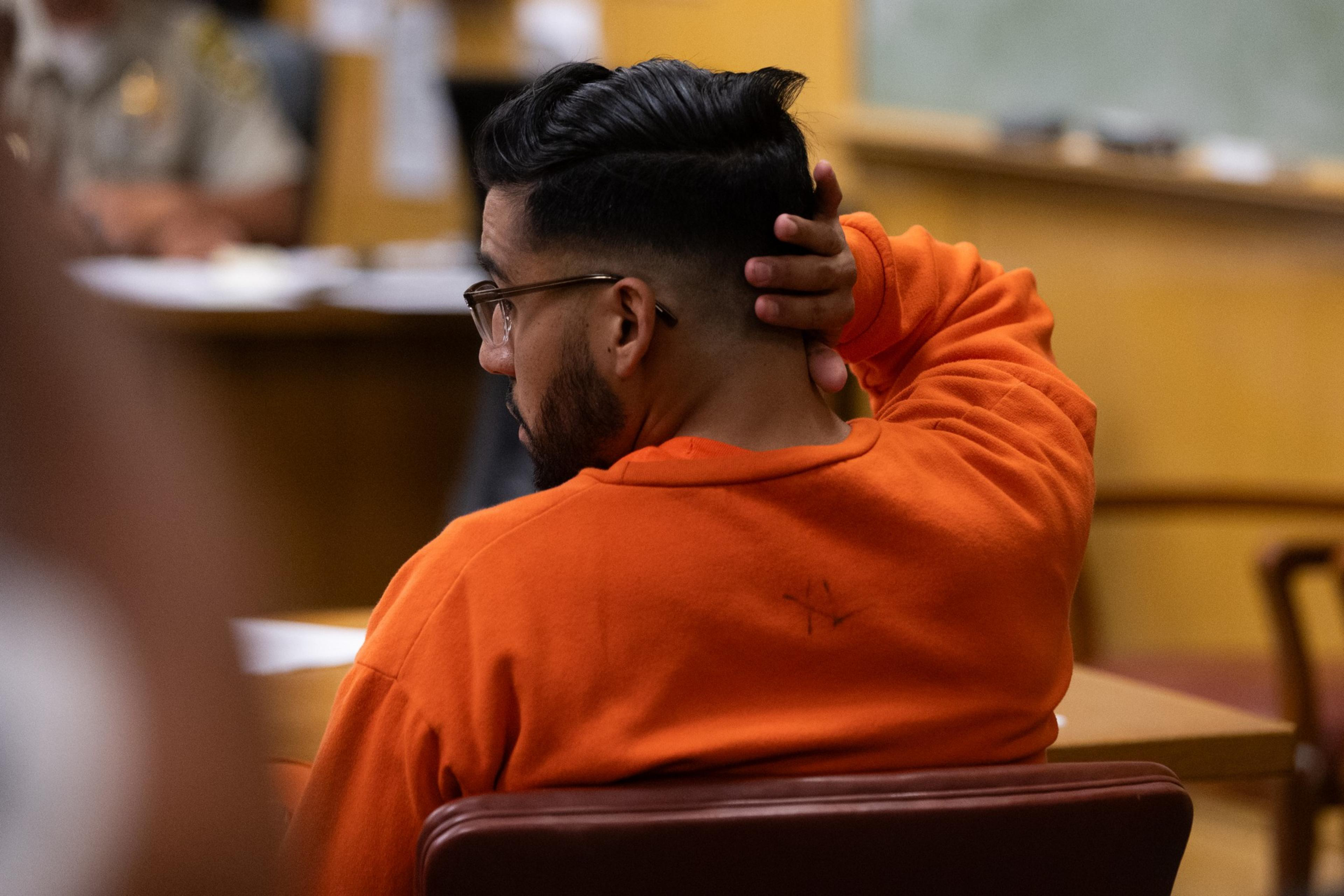 A bearded man with glasses in an orange top sits in a courtroom, facing forward with his hand on the back of his head.