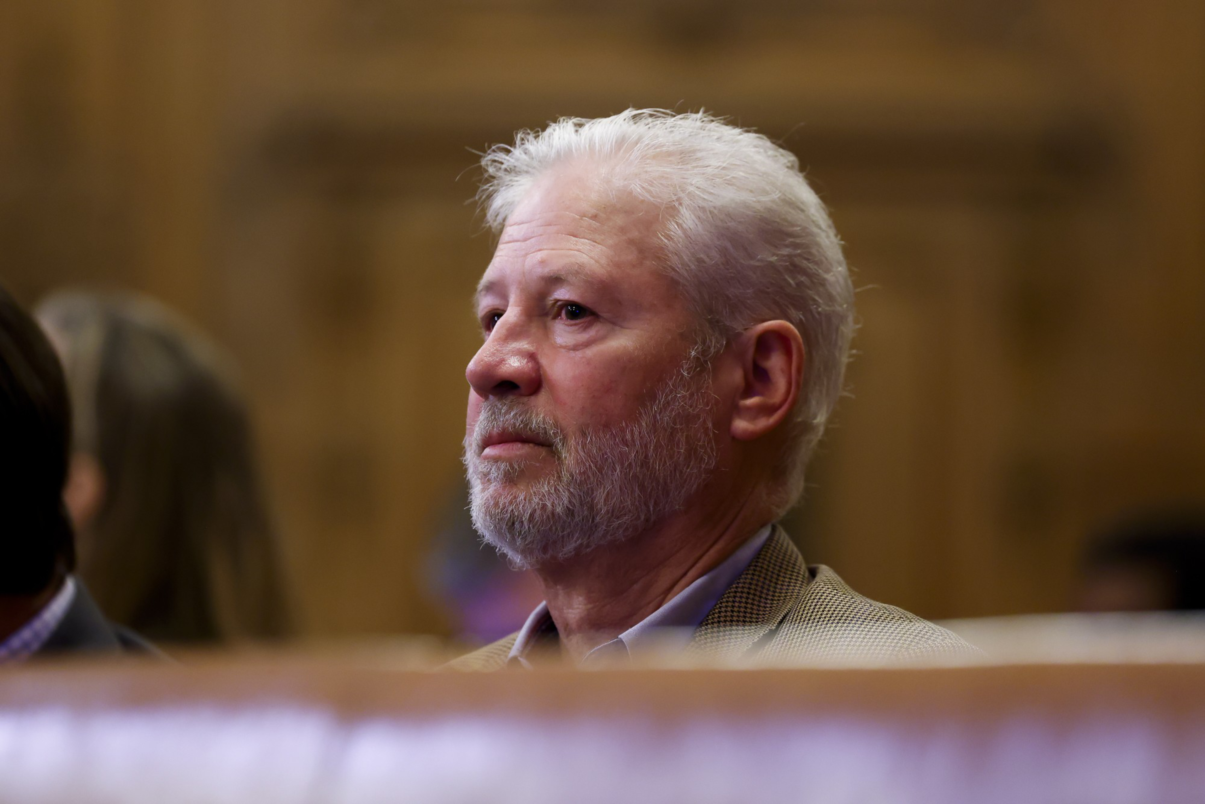 A man with white hair and a beard is seated, looking attentively forward. He's wearing a beige jacket and there's a blurred background behind him.