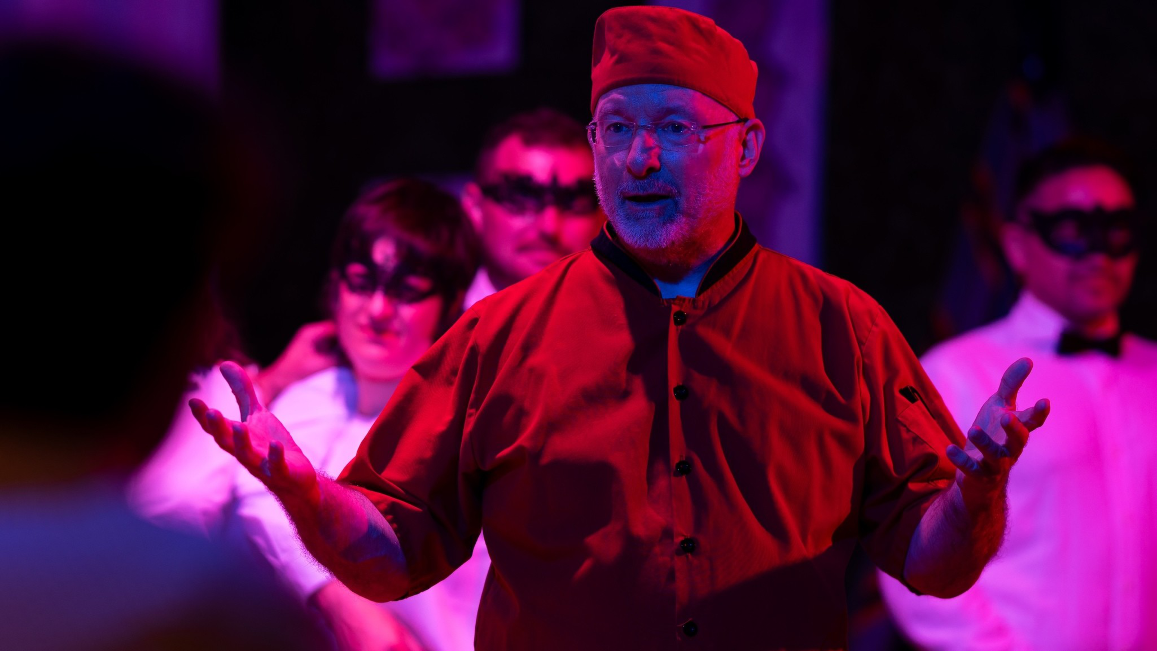 A man in a red shirt and hat speaks with open hands, while others in white shirts and black masks stand behind him in dim, colorful lighting.