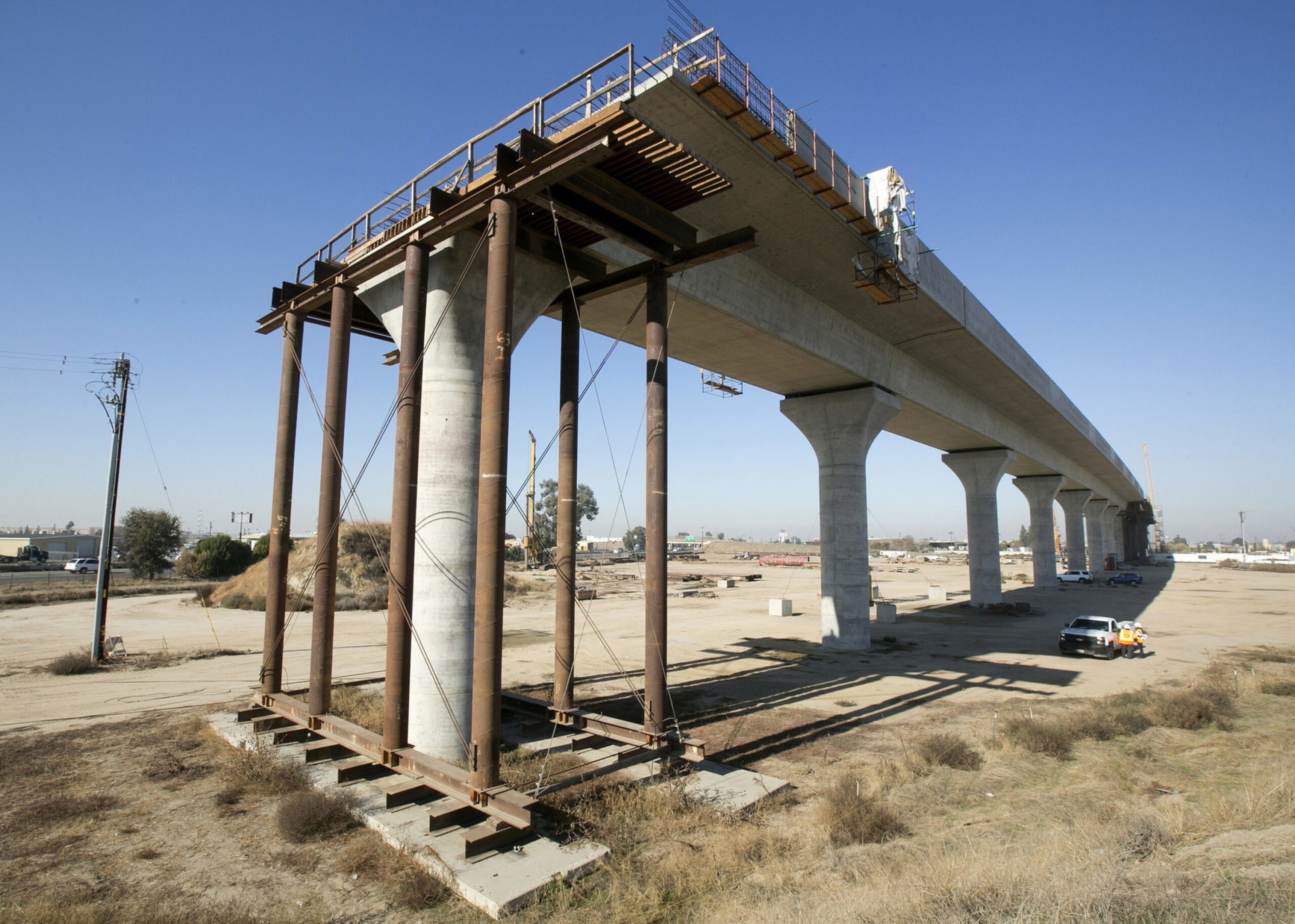 A partially constructed elevated concrete bridge spans across dry, open land with support columns and temporary metal scaffolding.