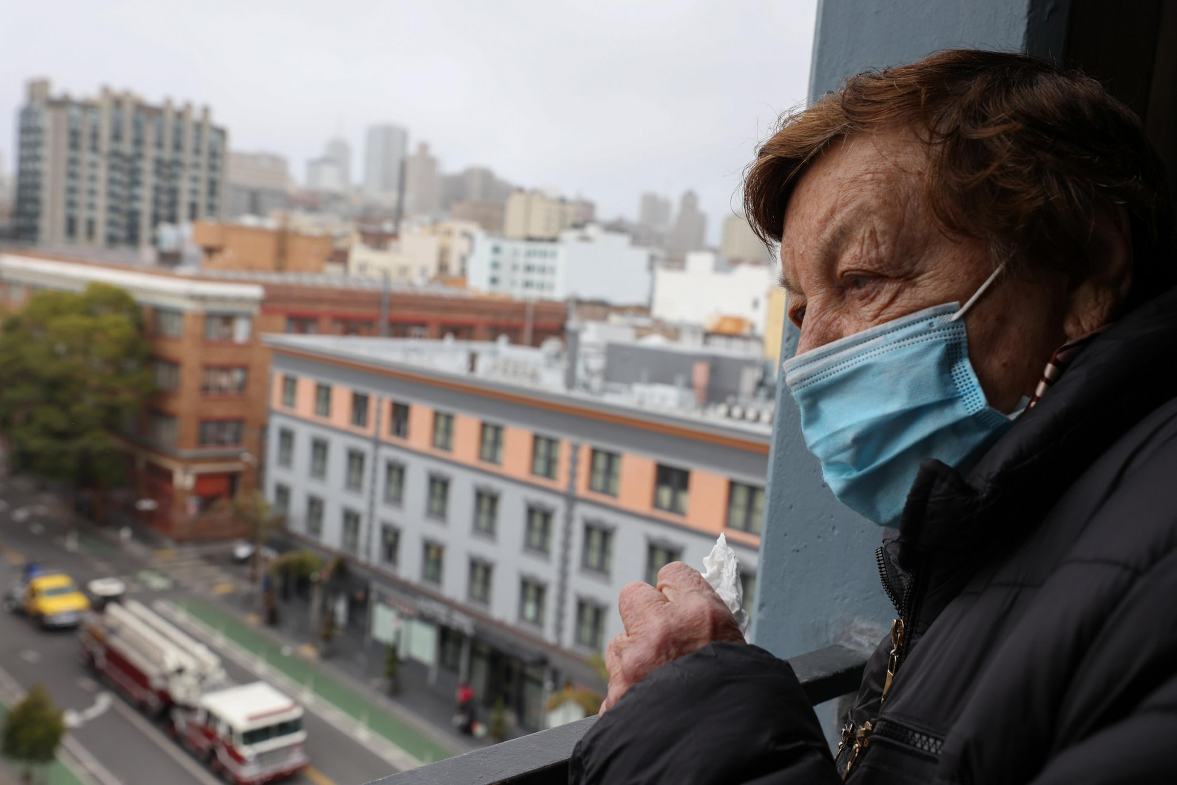 An elderly person wearing a blue surgical mask looks out from a balcony at a city street below, holding a crumpled tissue in one hand.