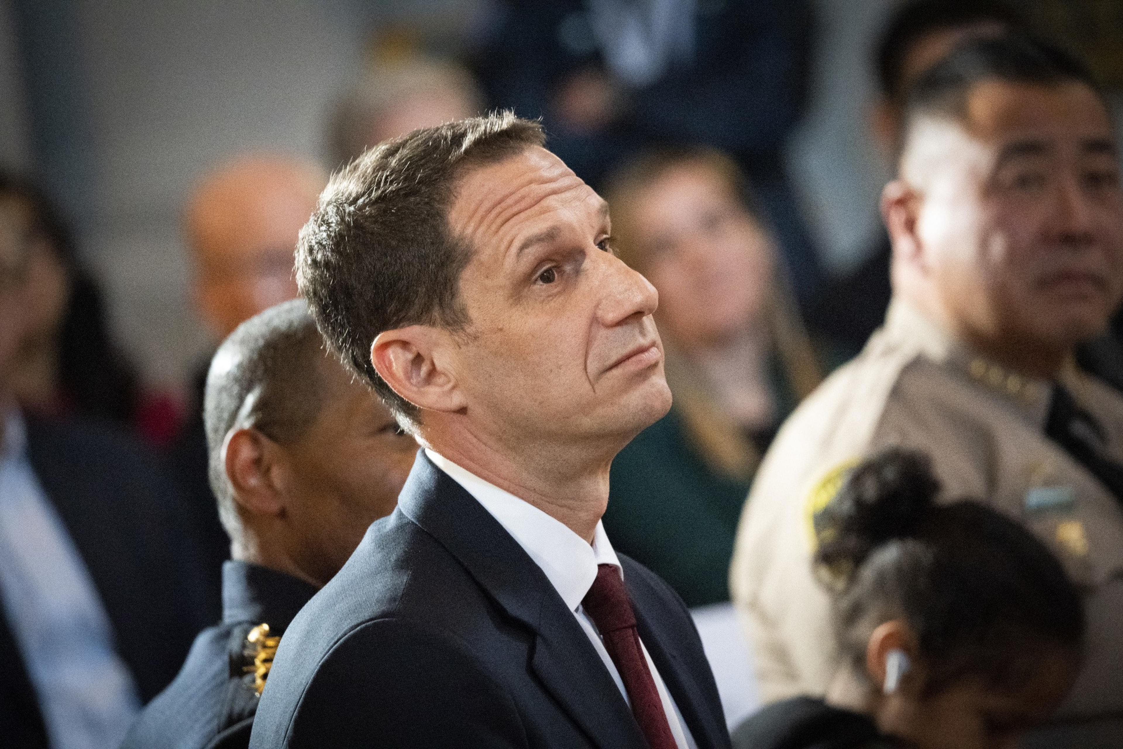 A man in a suit and tie looks upward, surrounded by people in formal attire, including a uniformed officer and a person in a tan suit. The setting appears formal.