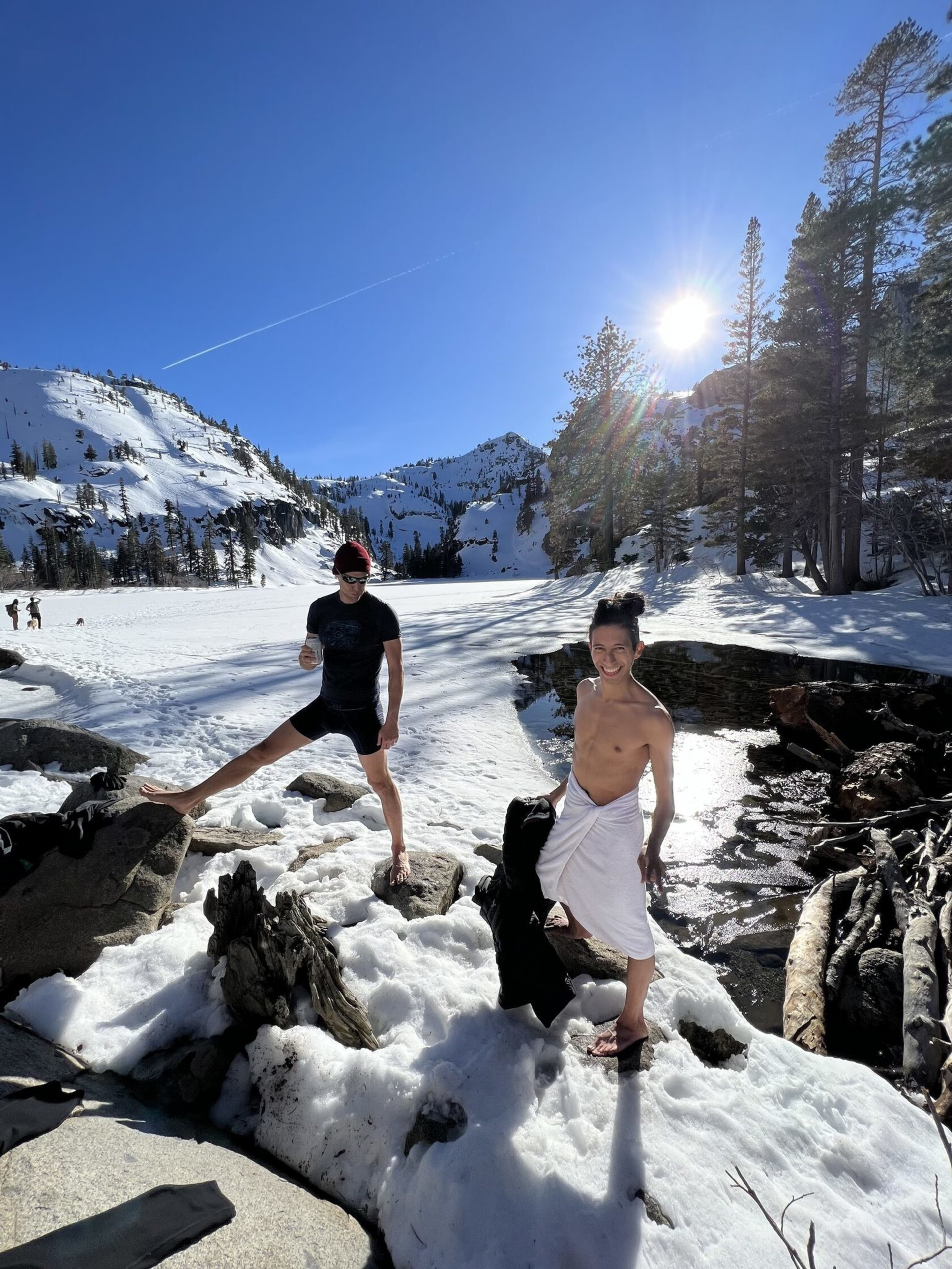 Two people stand on snow-covered rocks by a stream in a snowy mountain landscape. One wears a towel, the other sports shorts. The sun shines brightly above.