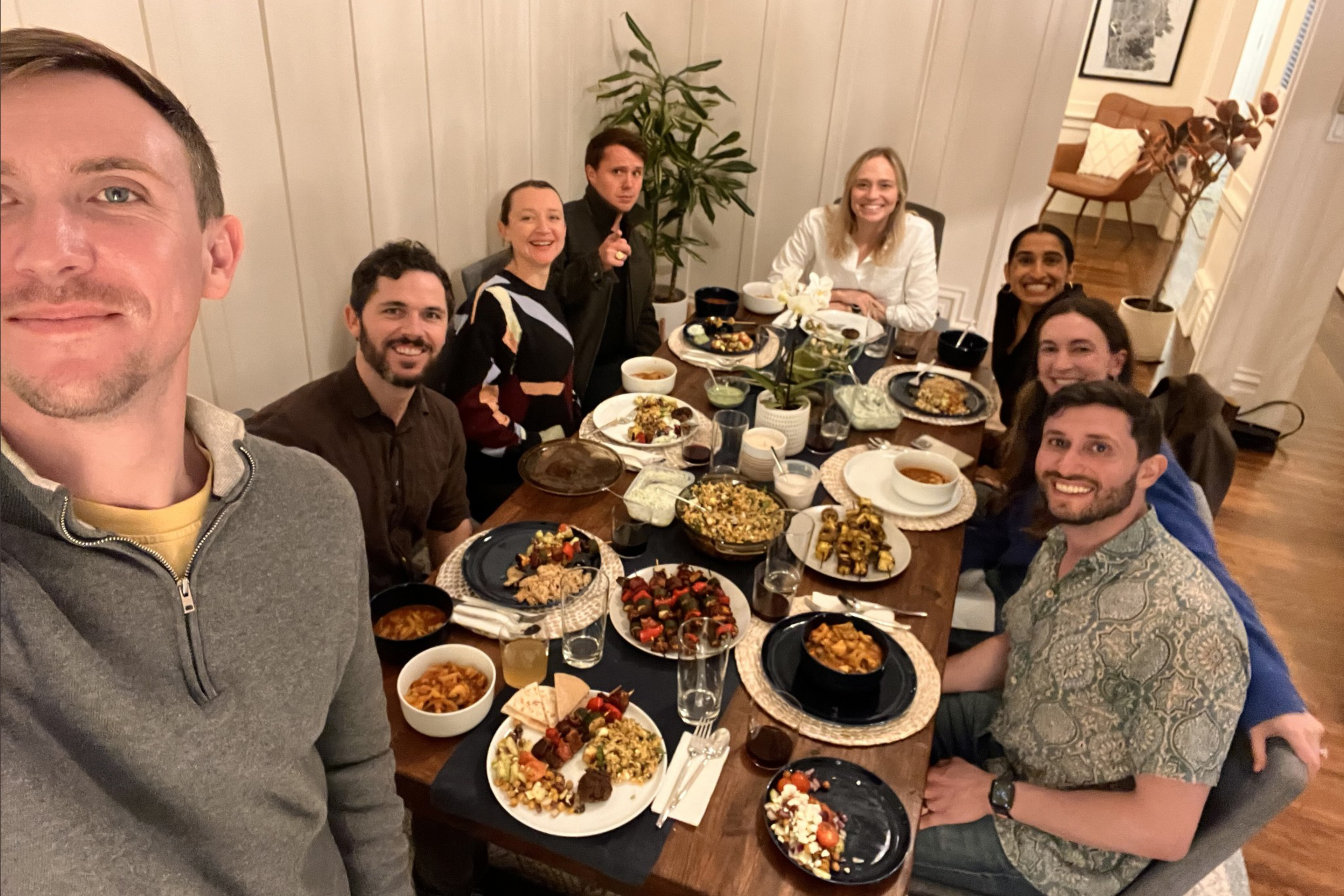 Eight people sit around a dining table filled with various dishes, smiling and enjoying a meal together in a cozy, warmly lit room.