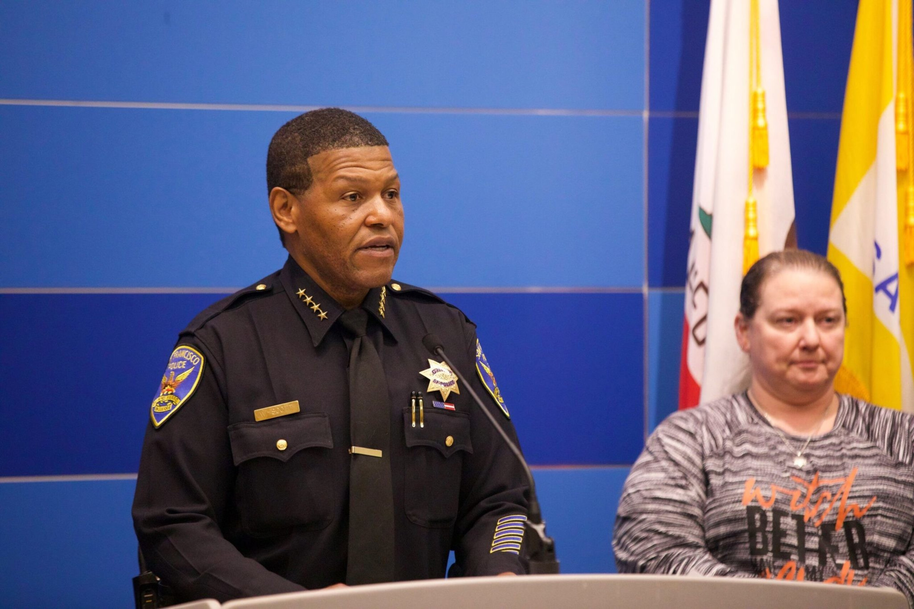 A man in a police uniform speaks at a podium, with a woman in a gray patterned shirt standing beside him against a blue background.