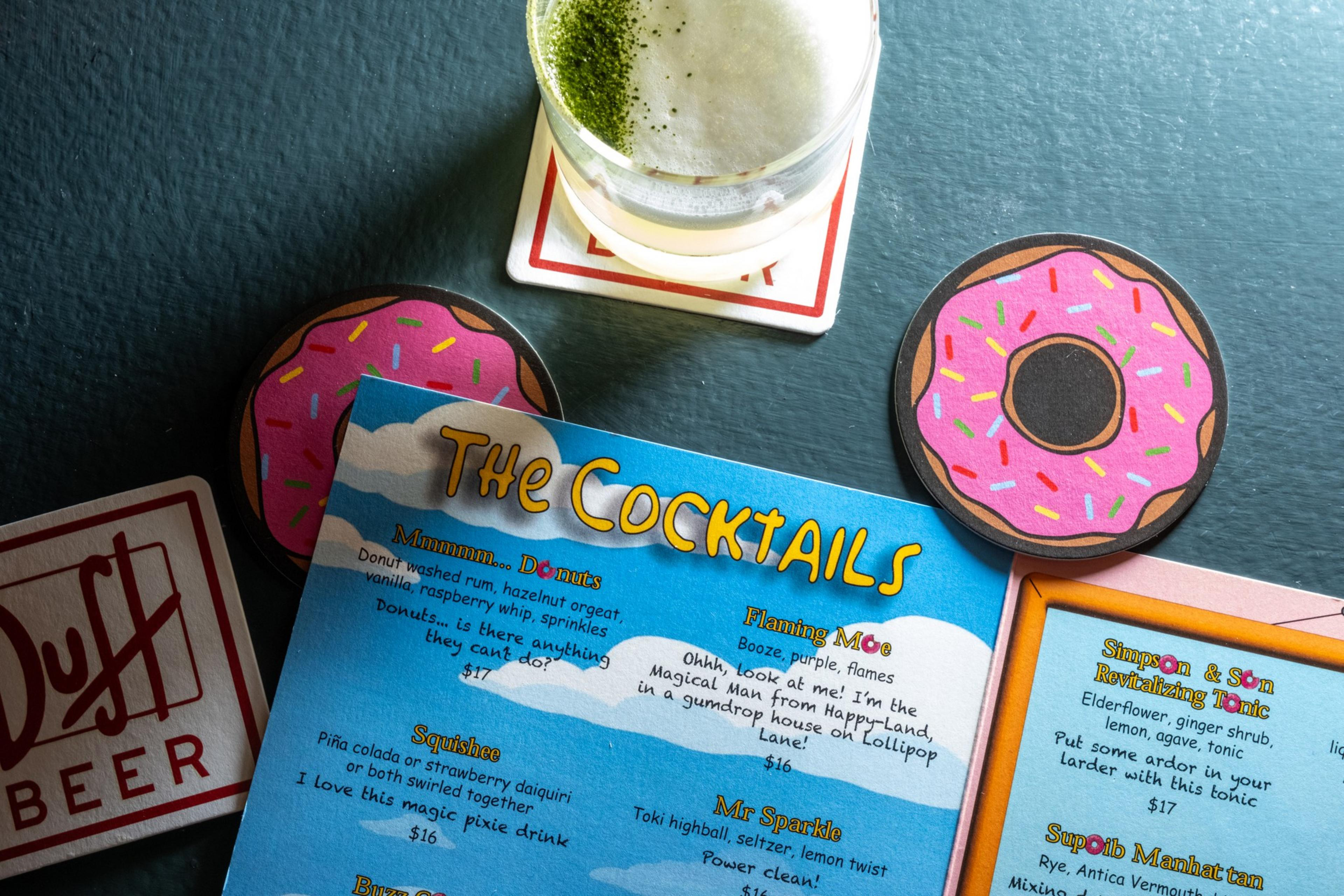 A cocktail menu featuring playful drink names is surrounded by donut-shaped coasters and a glass of a frothy green drink on a &quot;Duff Beer&quot; coaster.