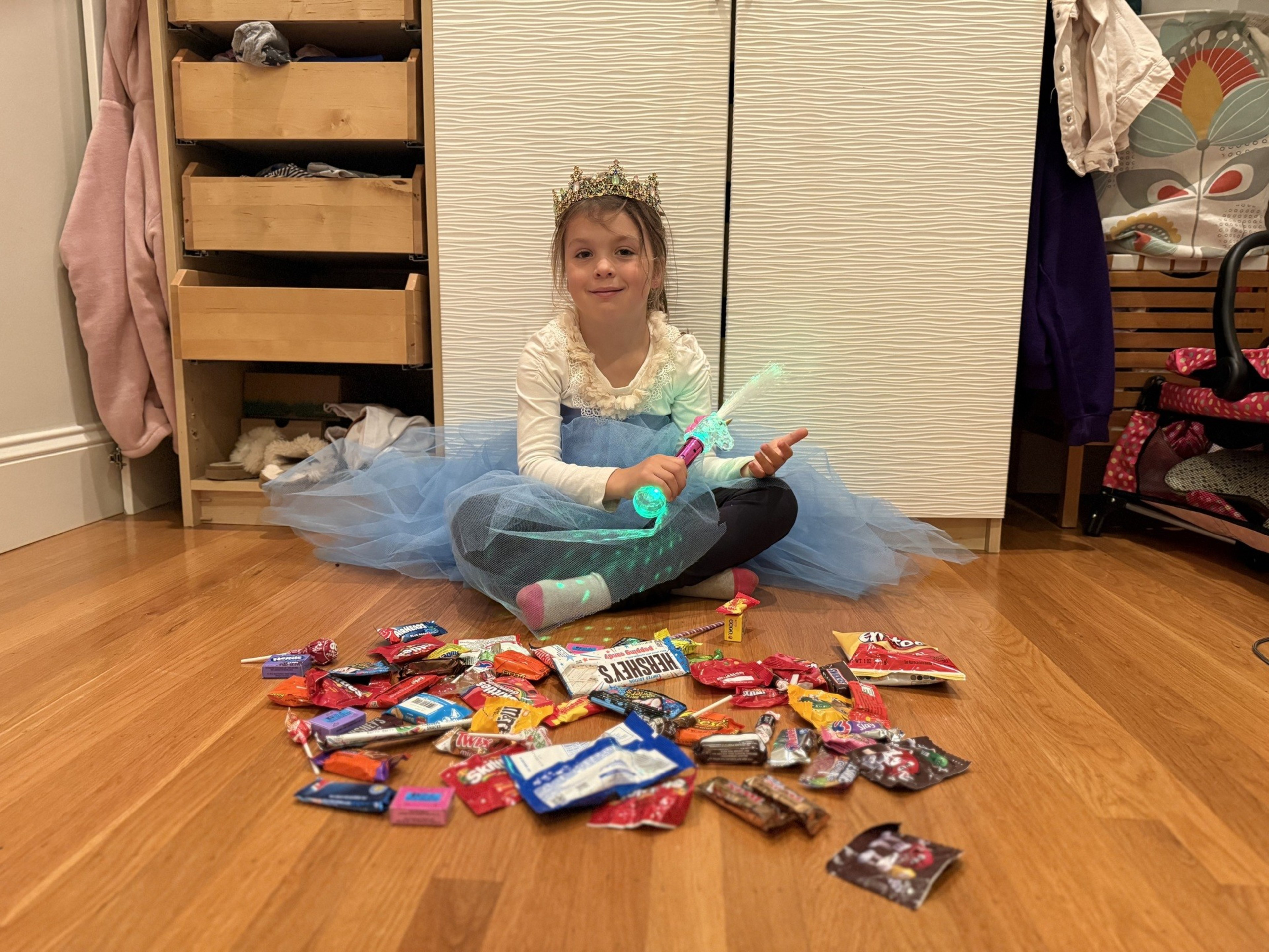 A kid in a blue dress with white long sleeves sits on the floor with multicolored candy in front of her.