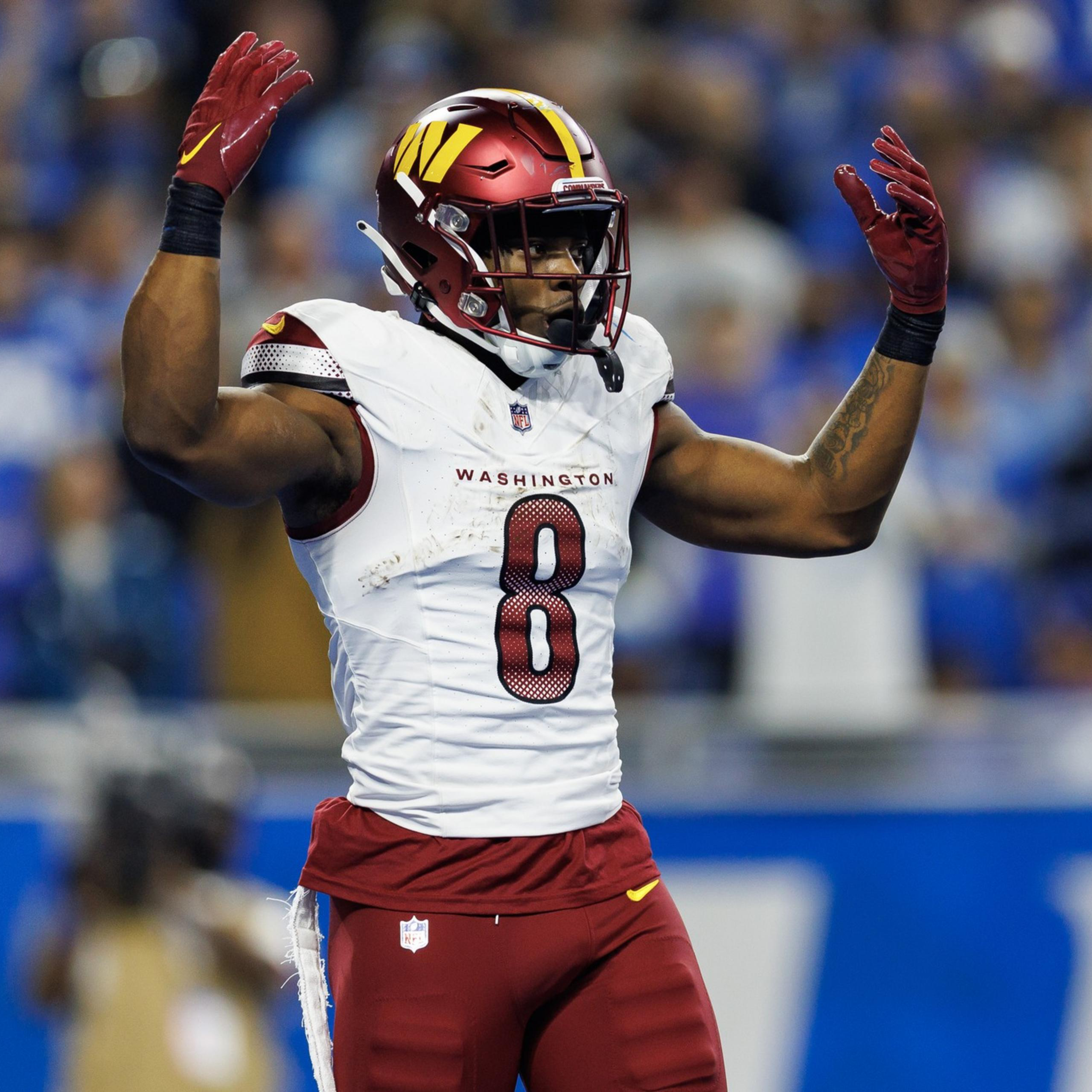 A football player wearing a Washington team uniform with the number 8 raises both arms, standing on a field with blurred spectators behind.