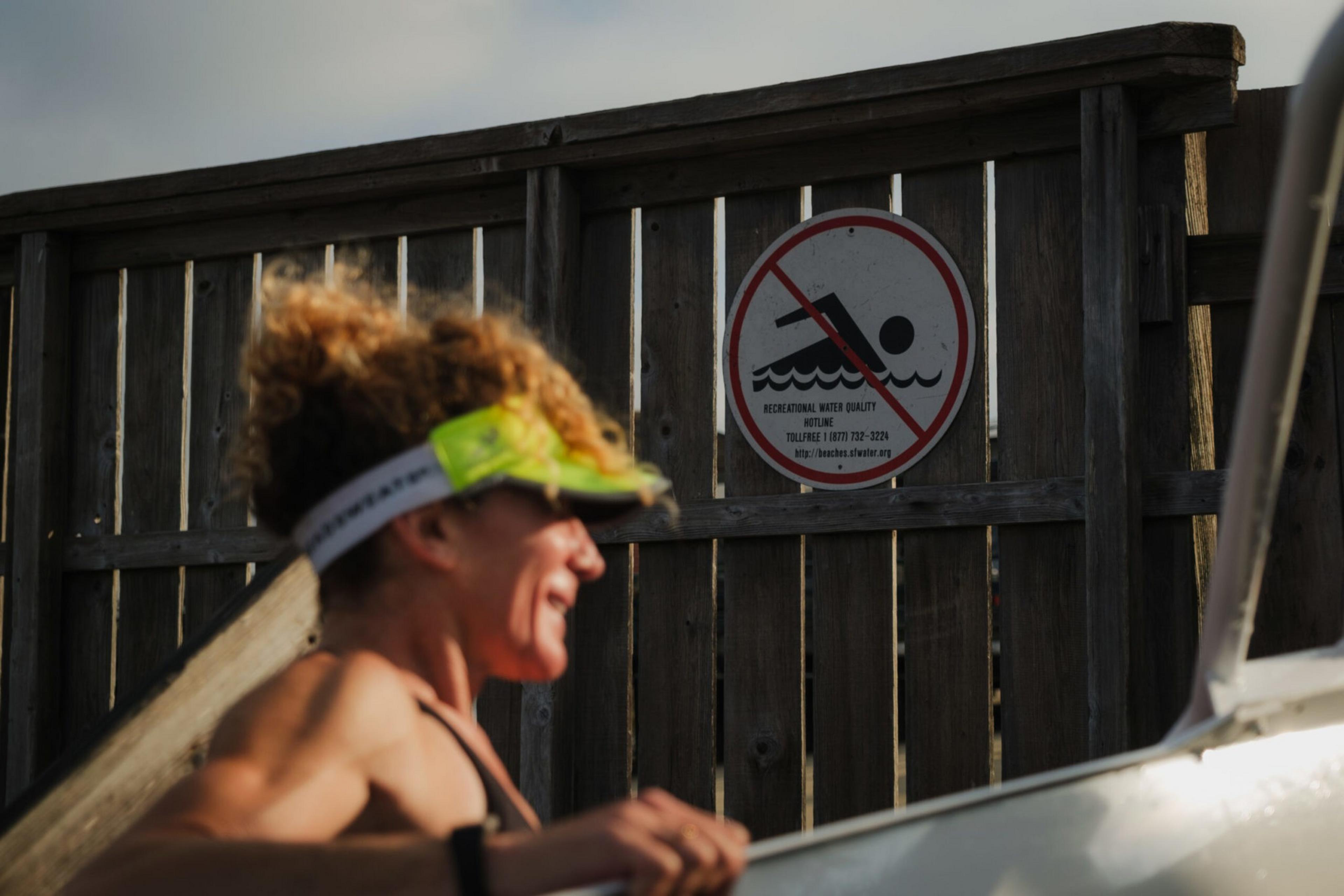 A smiling woman wearing a green visor stands near a wooden fence with a no swimming sign posted on it.