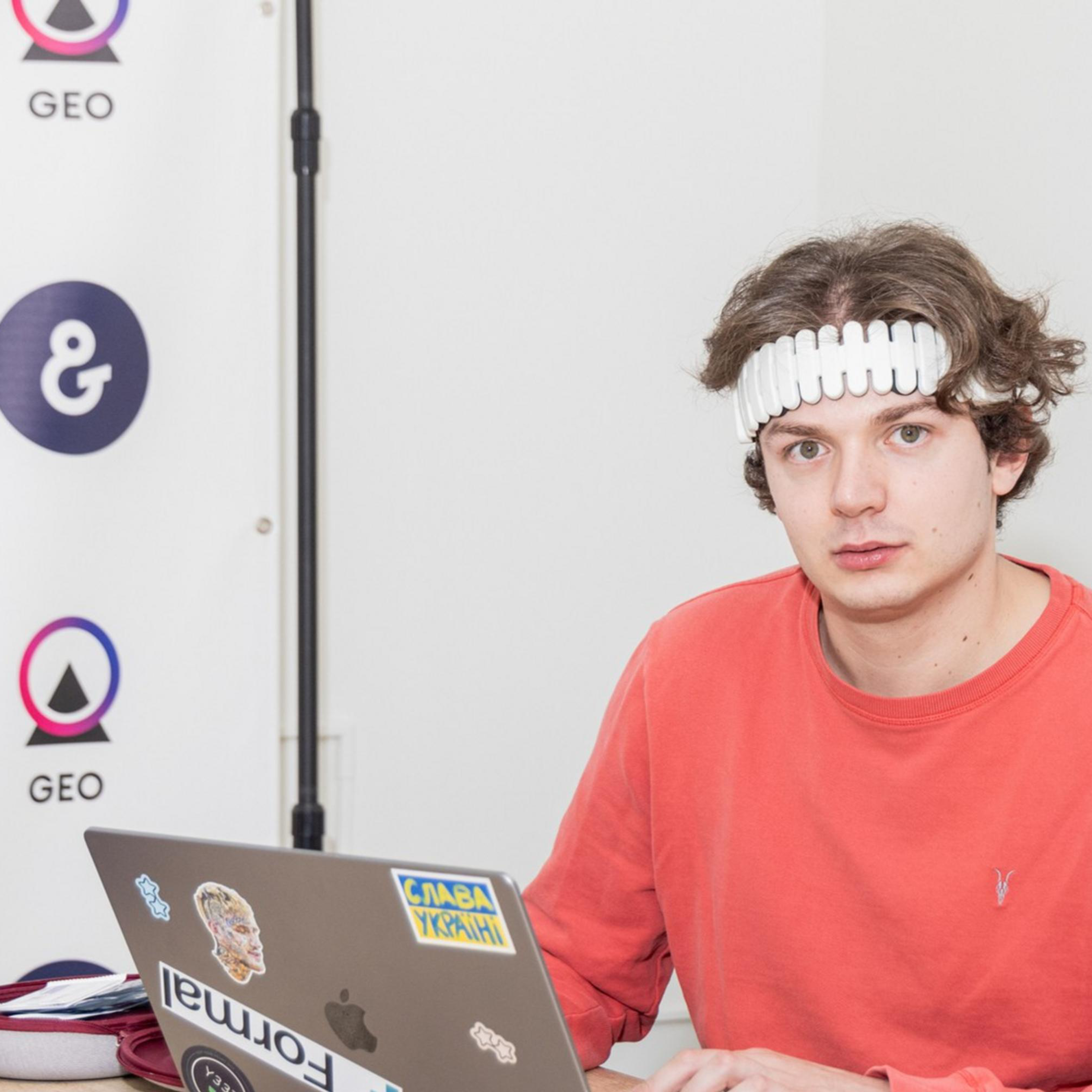 A person in a red shirt and a unique headband is sitting at a table using a laptop with various stickers. The background features simple white walls and logos.
