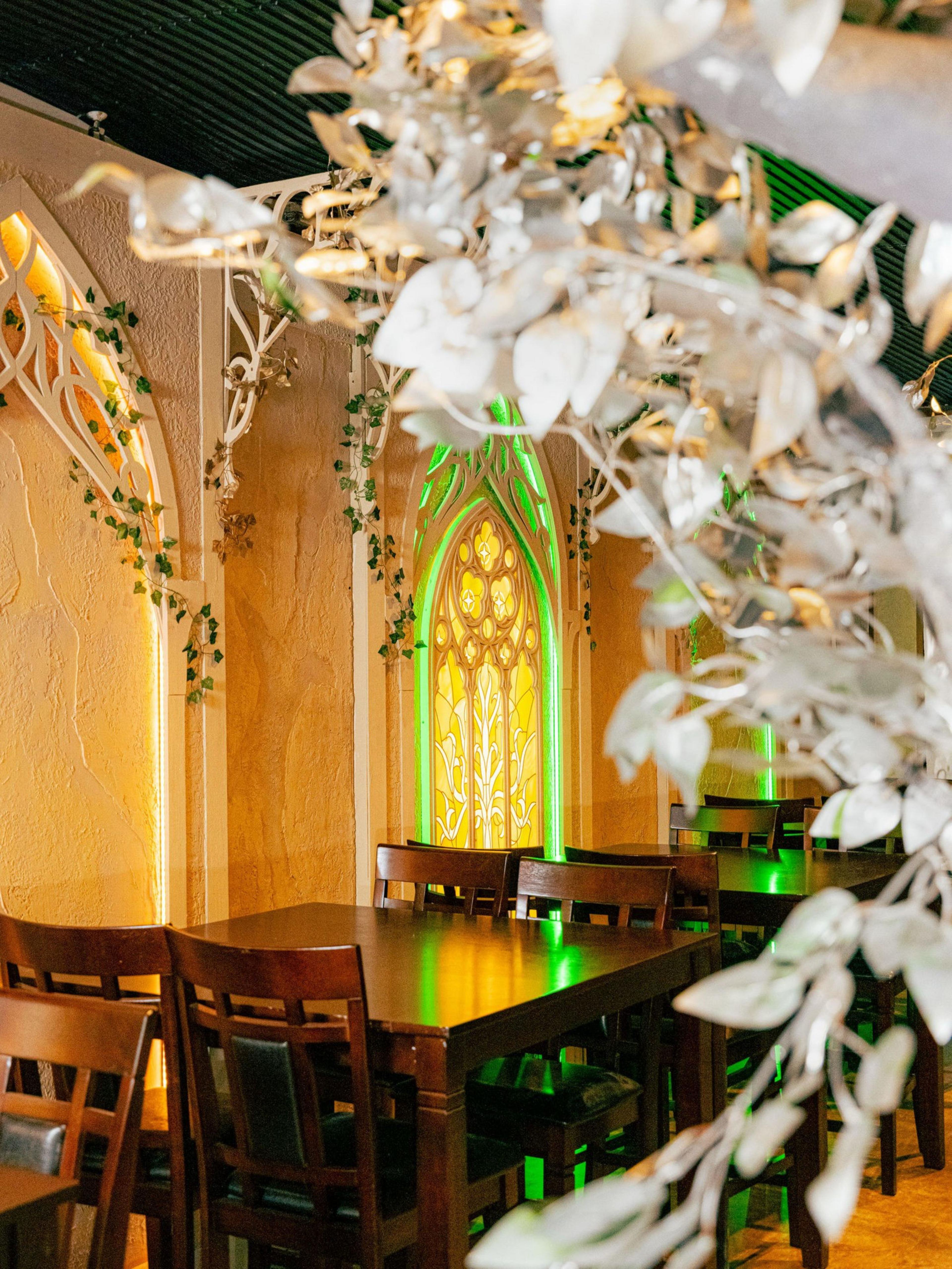 The image shows a cozy room with wooden tables and chairs, decorated with green and yellow stained glass windows, leafy vines, and warm lighting.