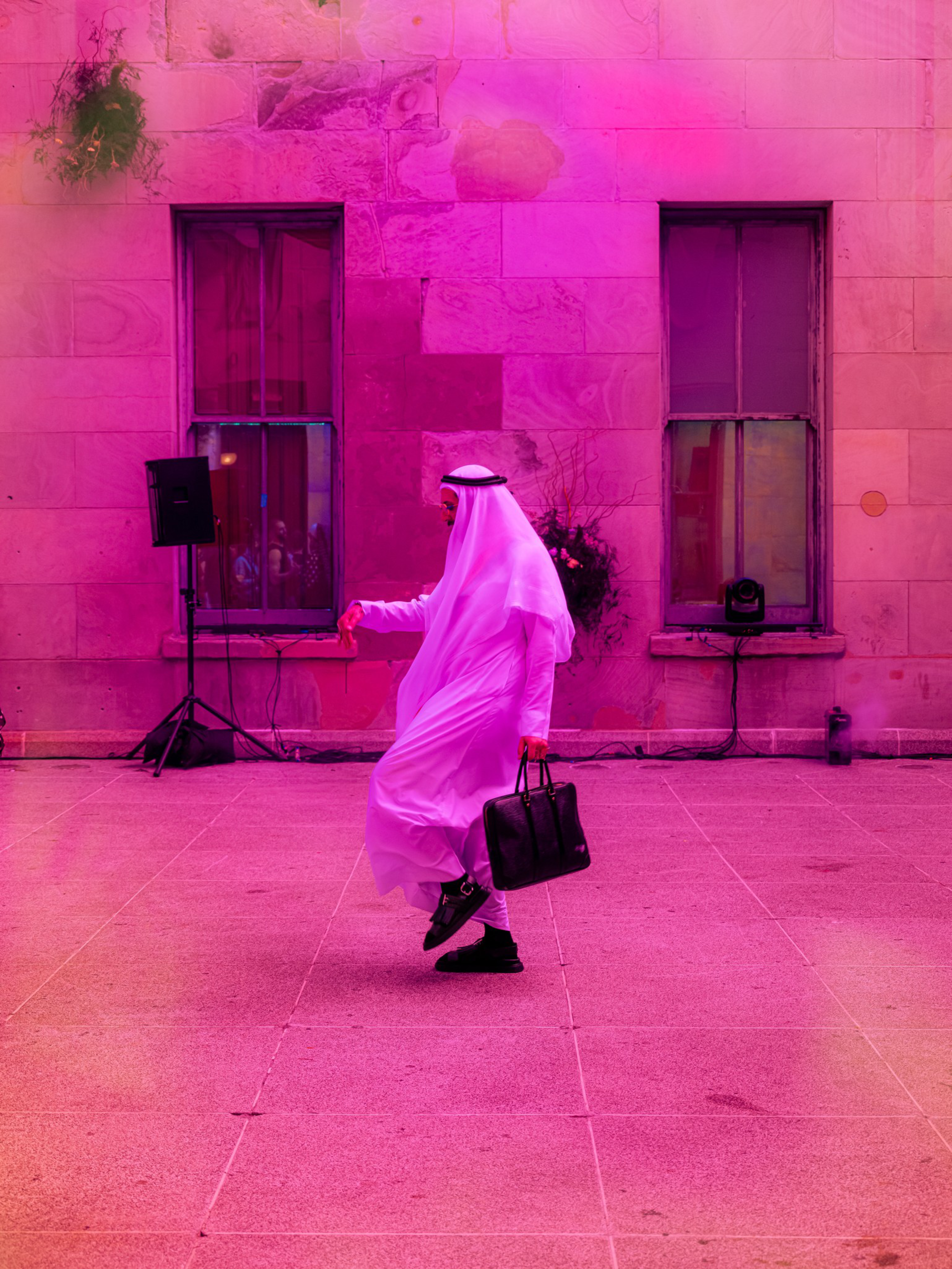 A person in traditional white attire and black shoes walks holding a black briefcase against a stone wall lit with pink and purple hues.