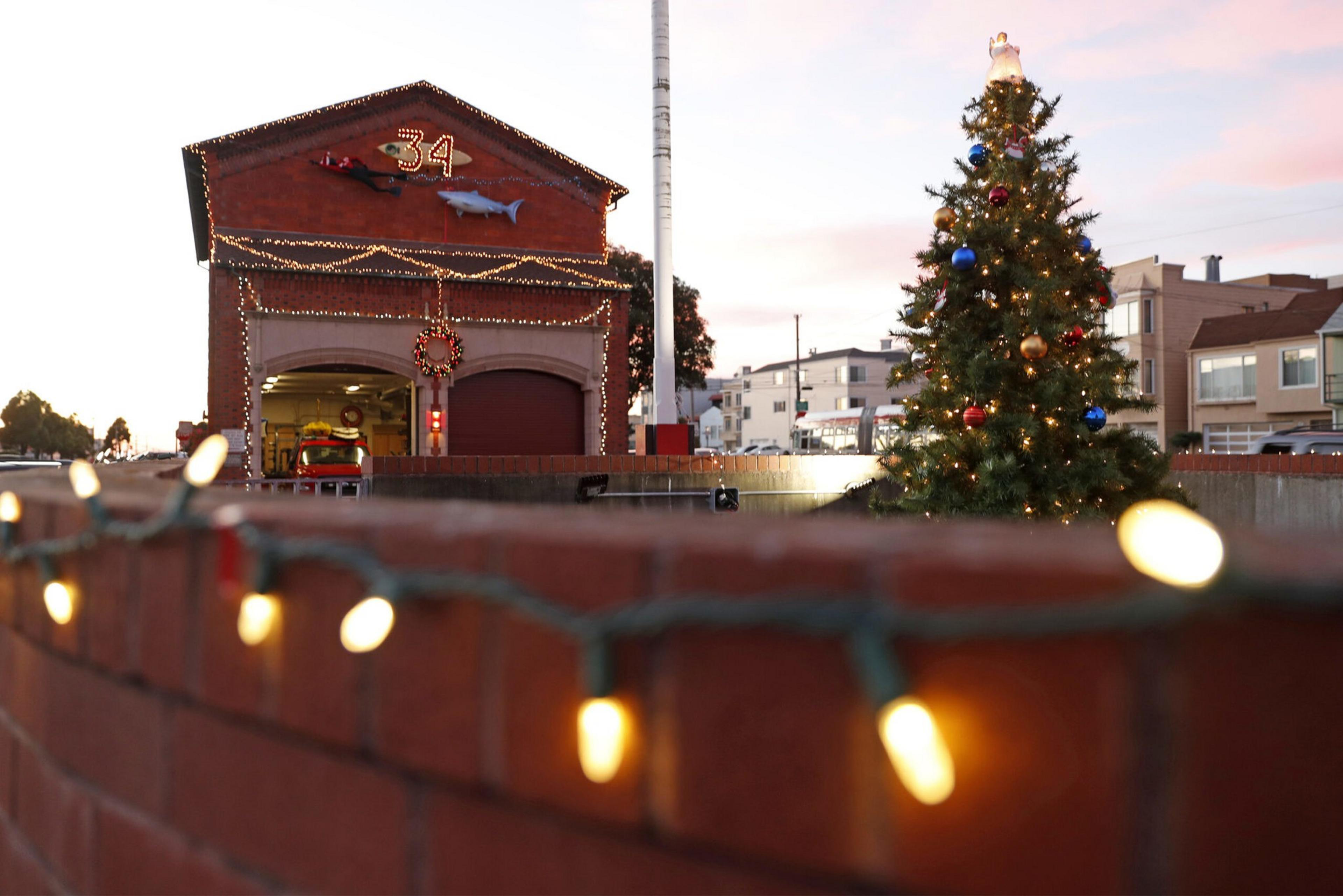 A fire station decorated with Christmas lights and a wreath, a large Christmas tree with ornaments, and string lights on a brick wall in the foreground.