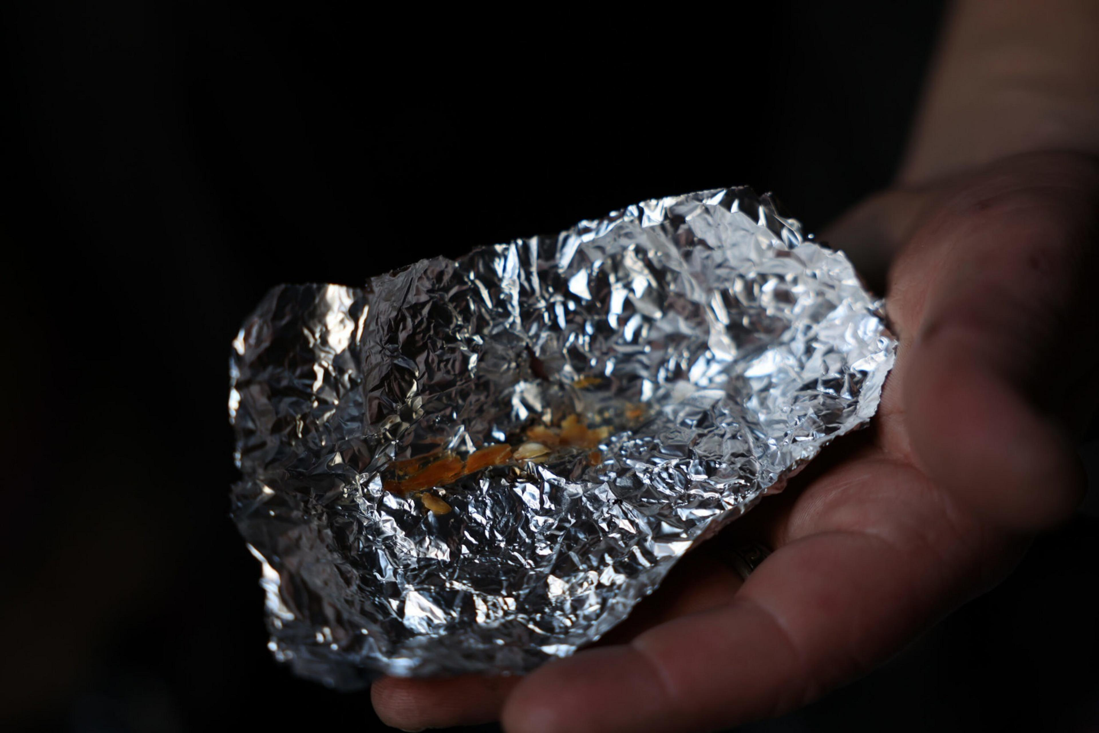 A hand holds a crumpled piece of aluminum foil with a small, orange residue inside, set against a black background.