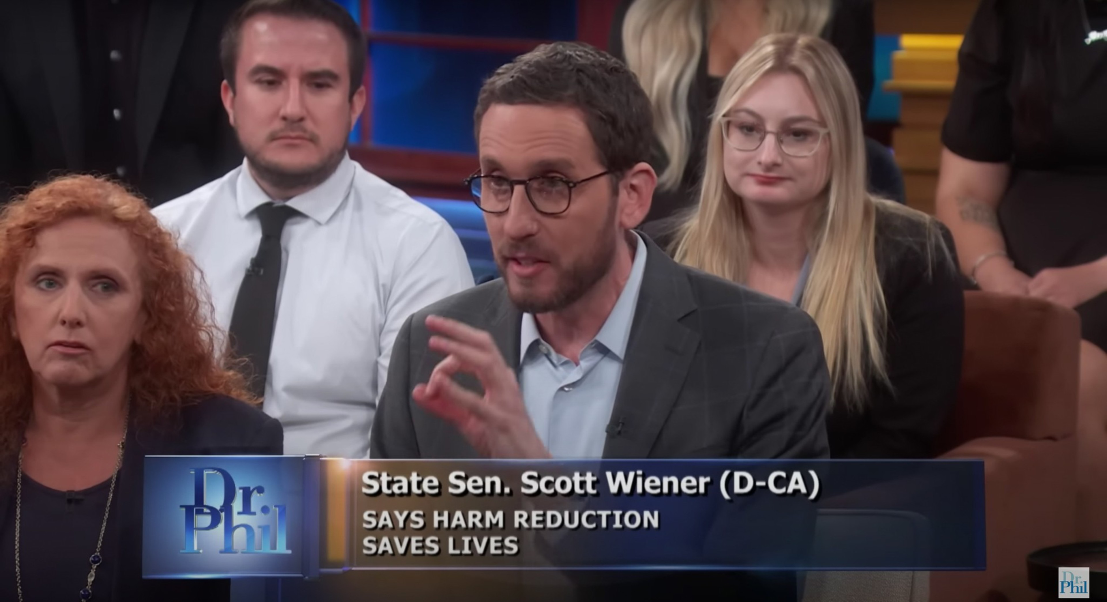 A man in glasses and a suit speaks passionately, with a group of attentive people behind him, and a caption about harm reduction on Dr. Phil.