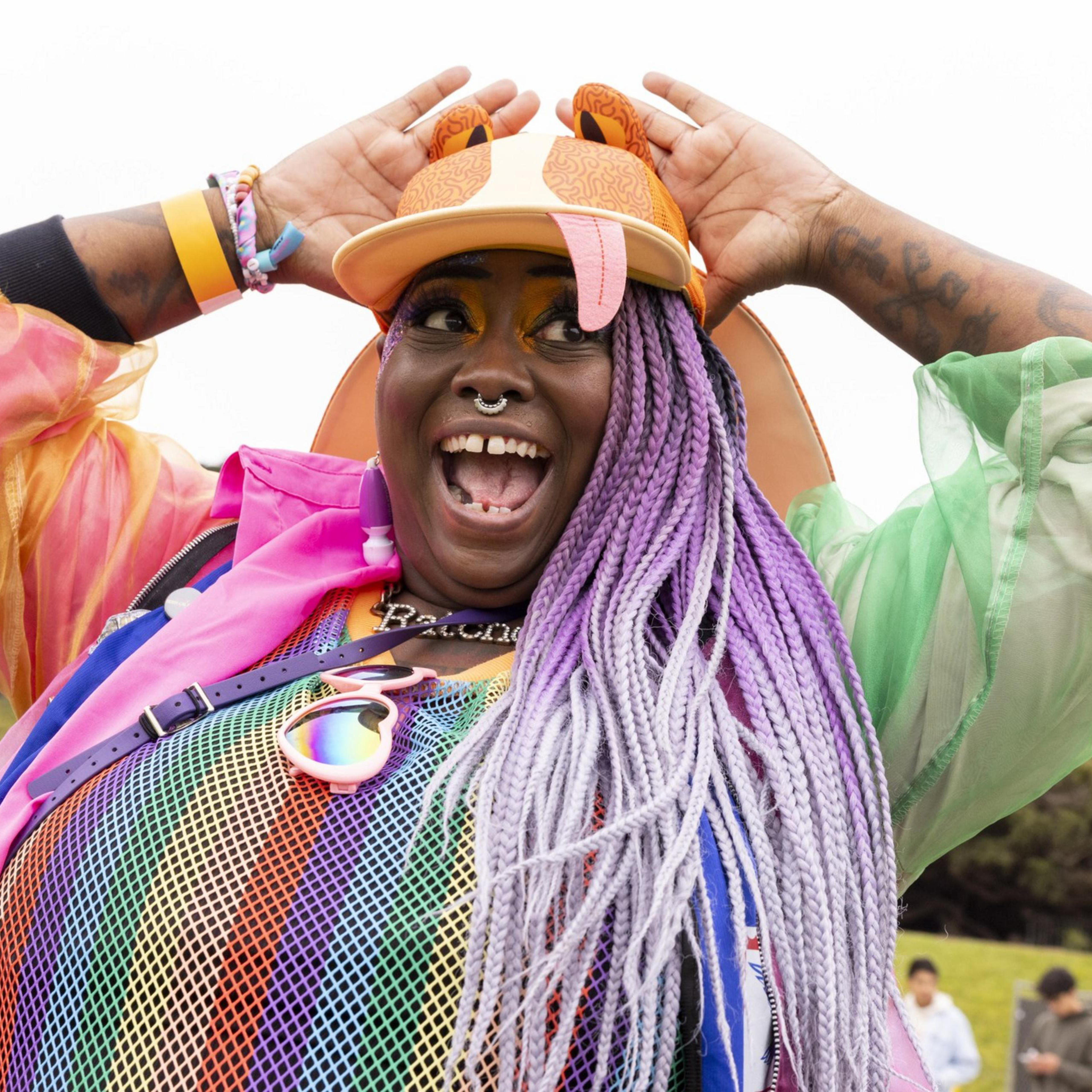 A joyful person with colorful braided hair and a vibrant rainbow outfit poses excitedly with hands on their hat, surrounded by an outdoor event setting.