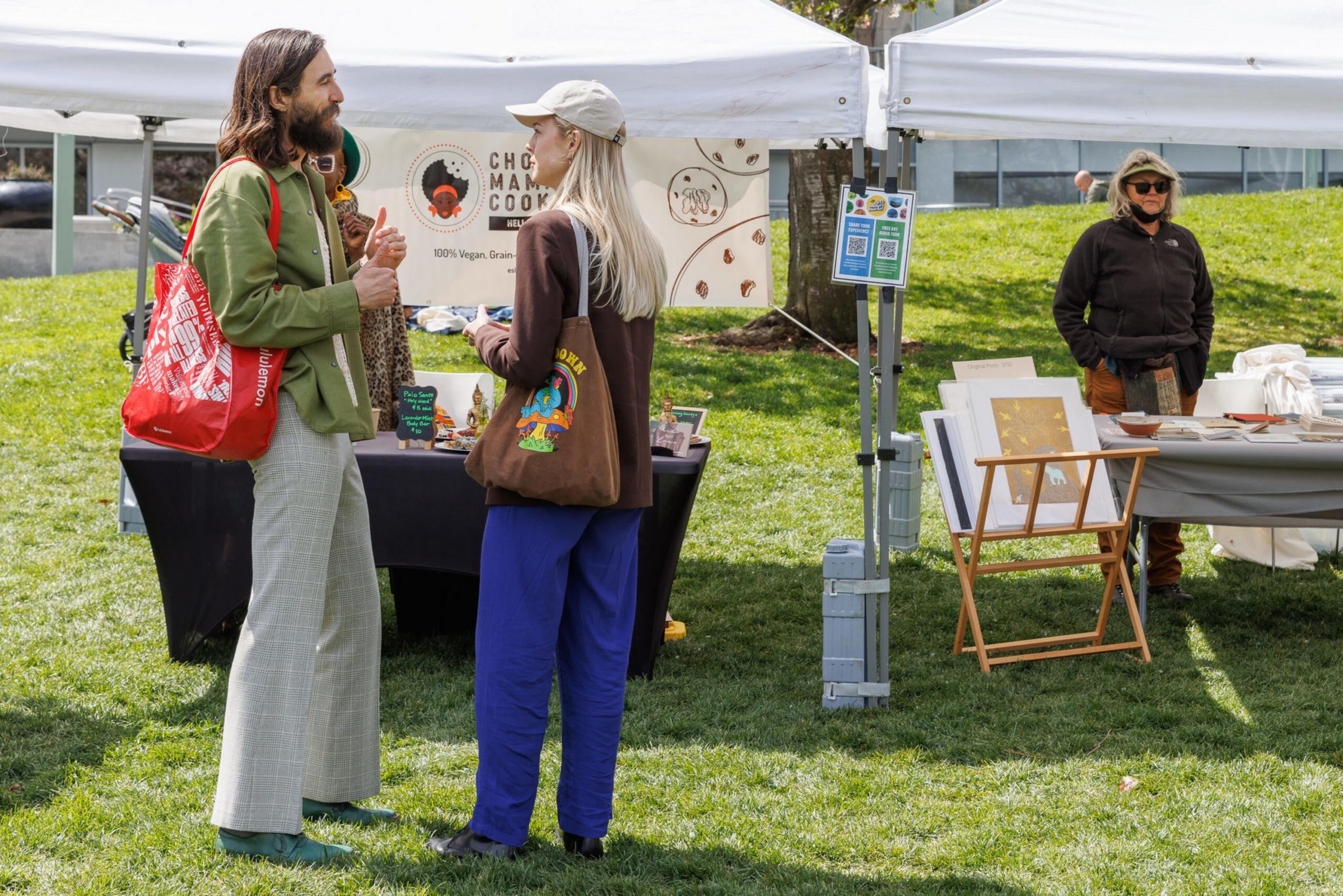 Two people chat at an outdoor market stall with cookies on display.