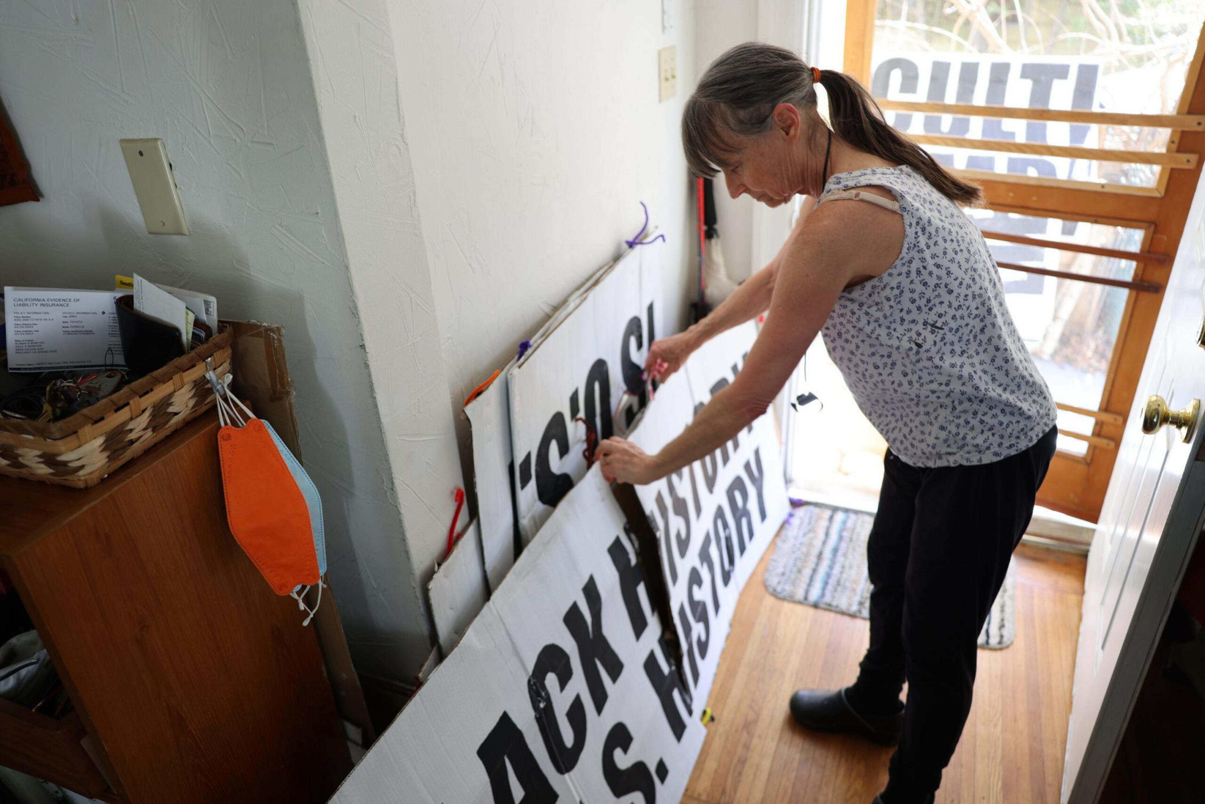 A woman with a ponytail adjusts large protest signs leaning against a wall inside a room with wooden flooring and a glass door.