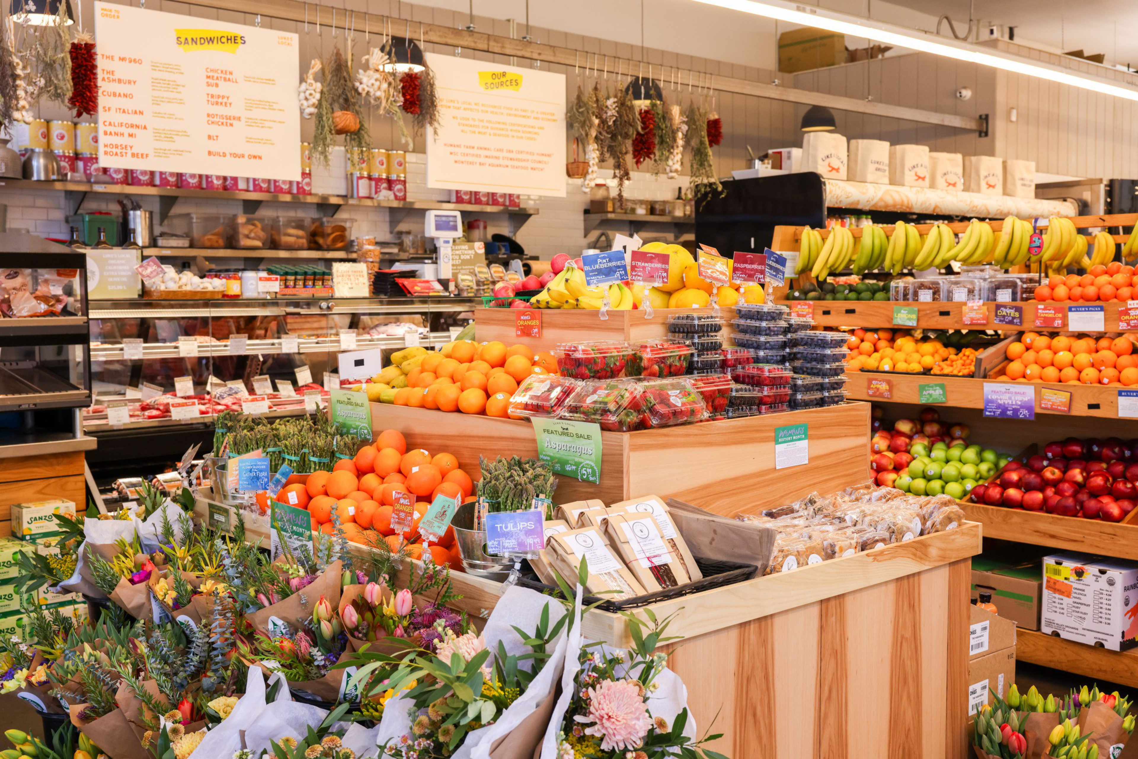 The image shows a colorful grocery store with fresh fruits like oranges, bananas, and berries displayed. There are flowers, asparagus, and a sandwich menu visible.