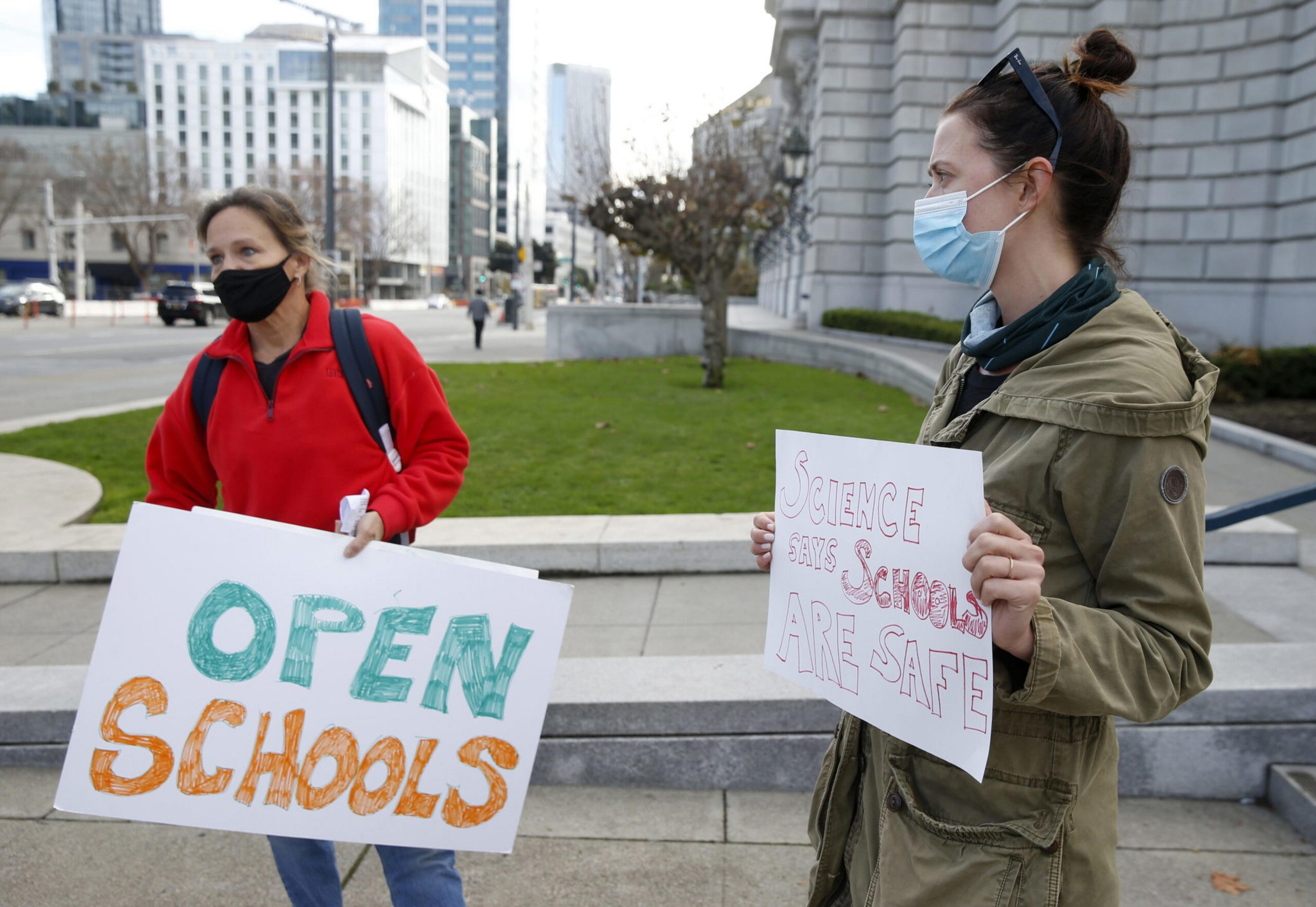 Two masked women hold signs advocating for open schools, with one sign reading "OPEN SCHOOLS" and the other "SCIENCE SAYS SCHOOLS ARE SAFE."