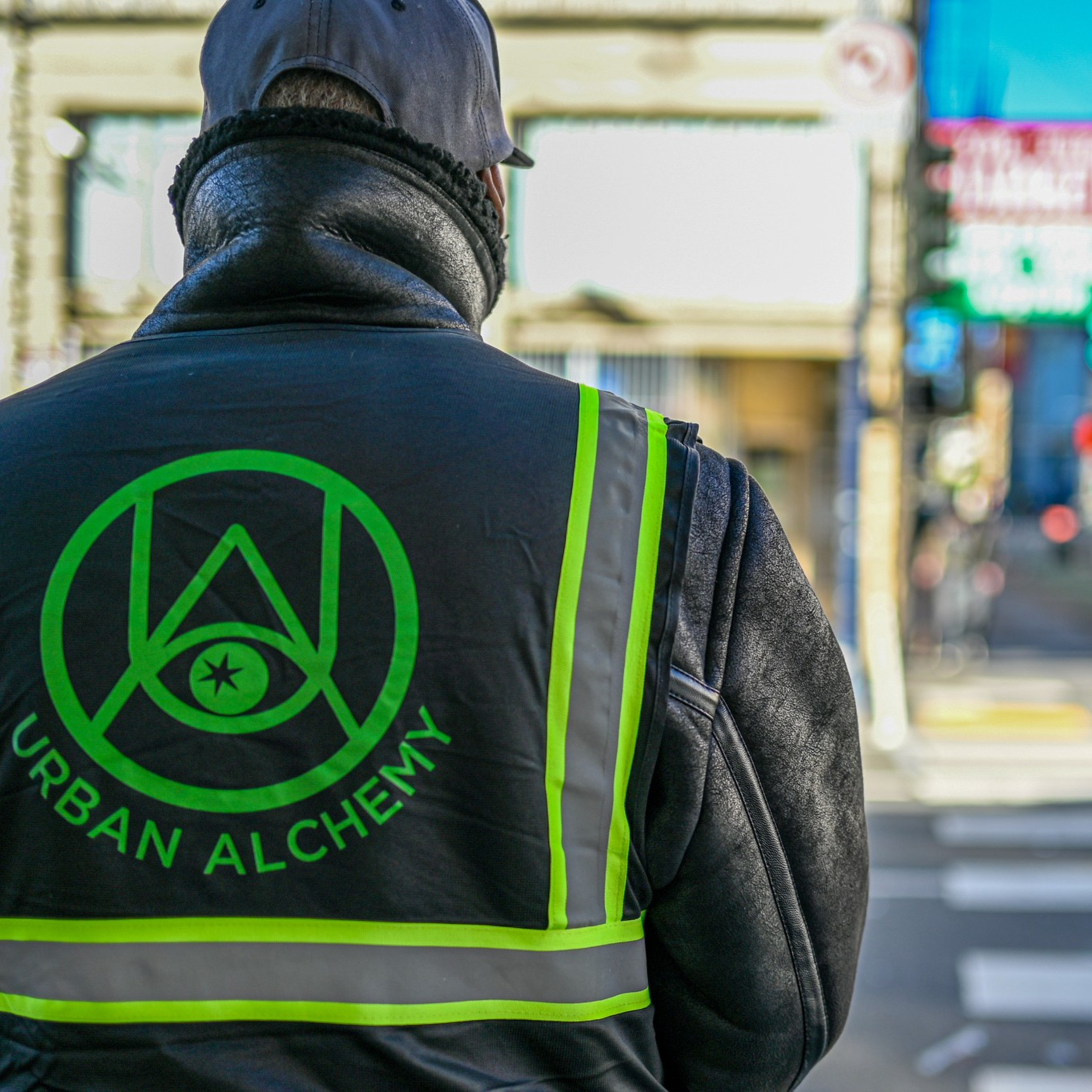 A person wearing a reflective vest with the words "Urban Alchemy" stands at a crosswalk, facing away from the camera in a busy urban street setting.