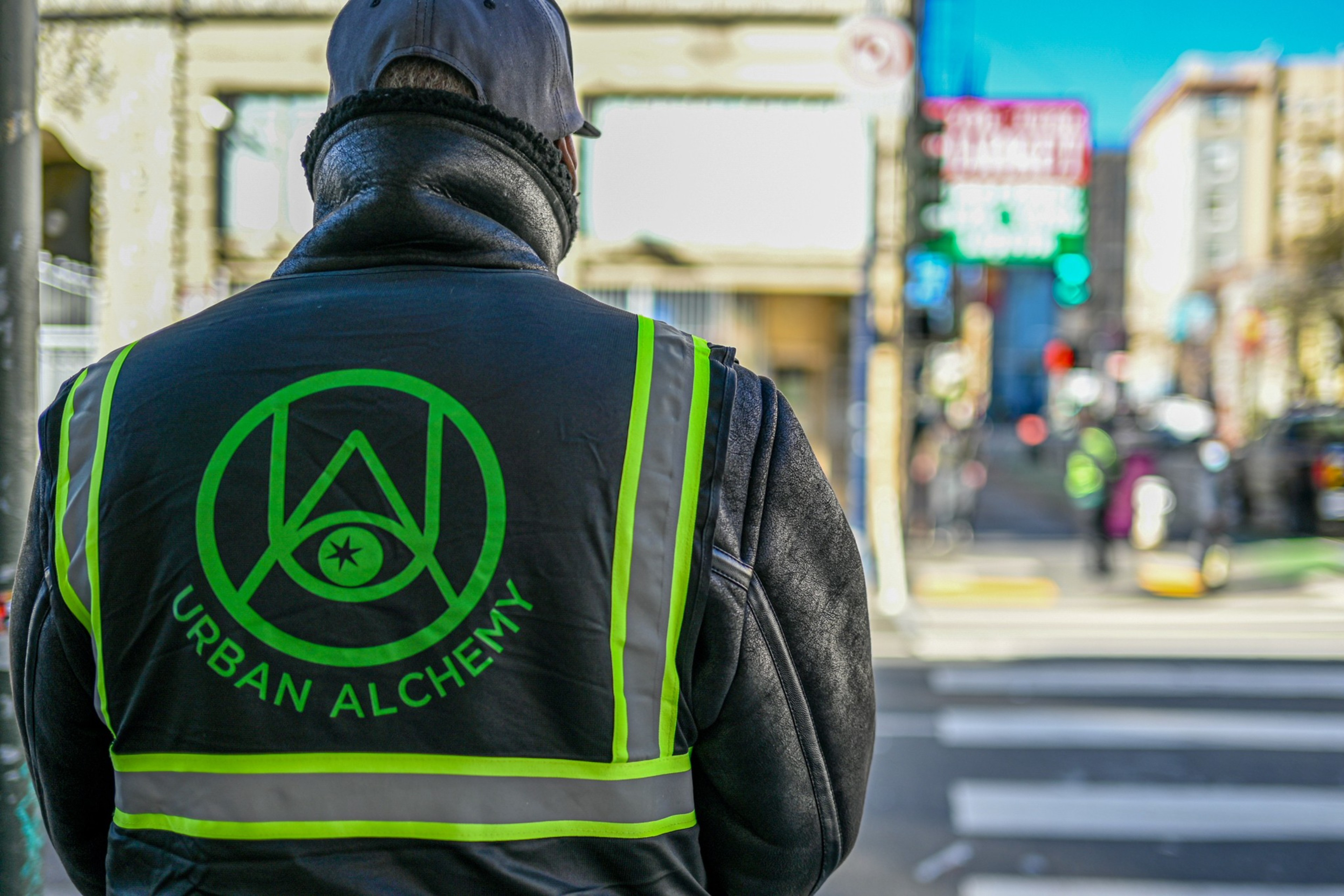 A person wearing a reflective vest with the words "Urban Alchemy" stands at a crosswalk, facing away from the camera in a busy urban street setting.