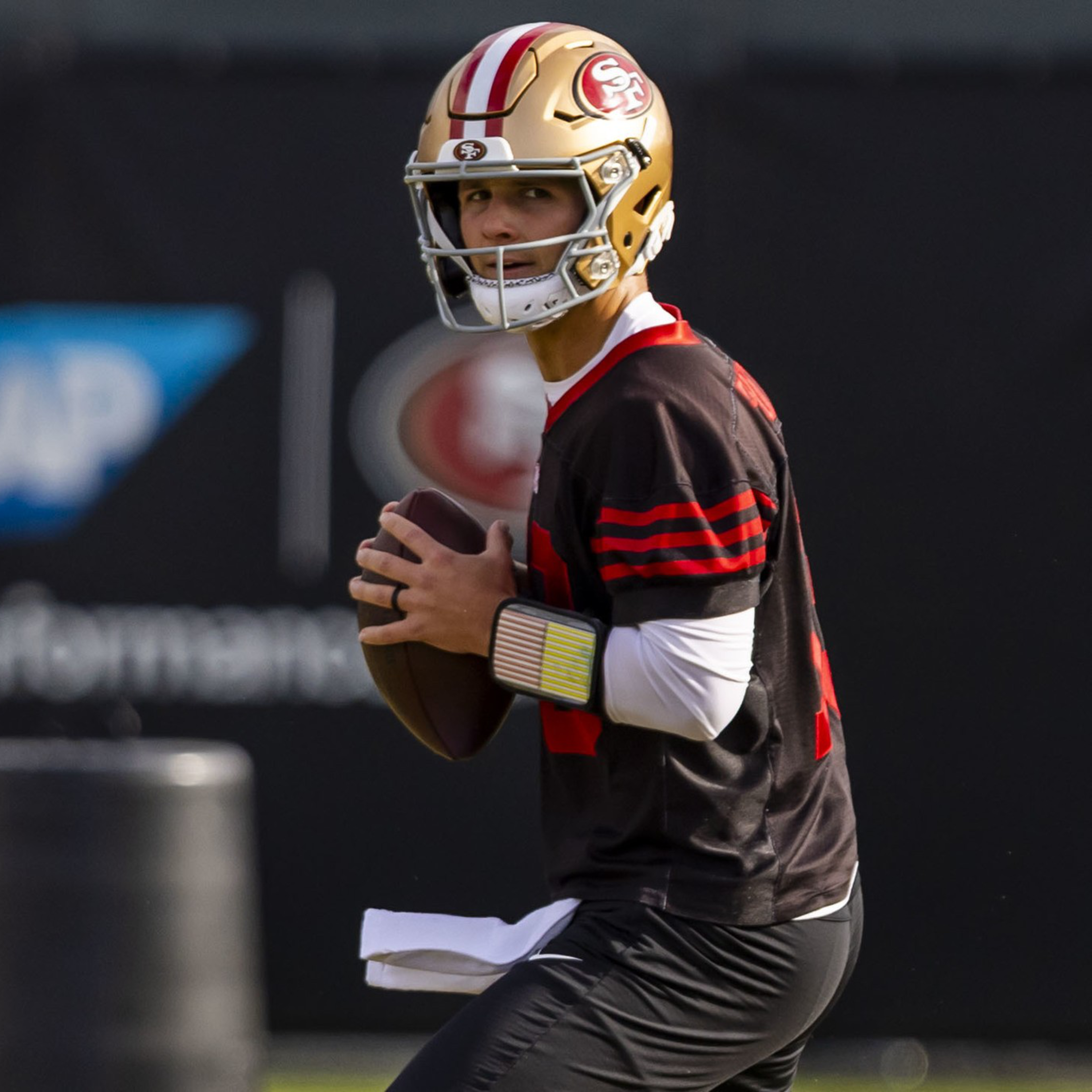A football player in a San Francisco 49ers helmet and uniform holds a football, preparing to throw during a practice session.