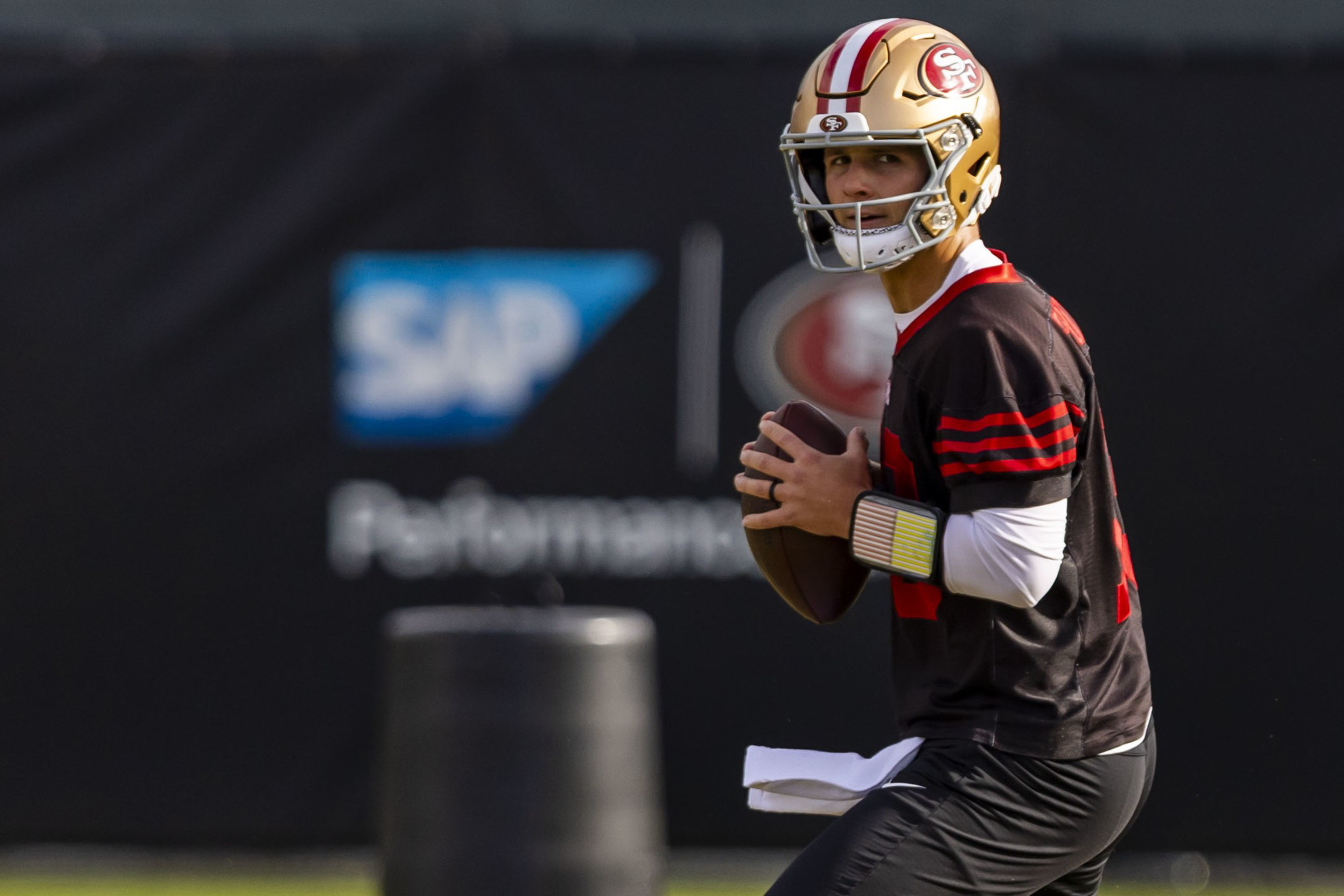 A football player in a San Francisco 49ers helmet and uniform holds a football, preparing to throw during a practice session.