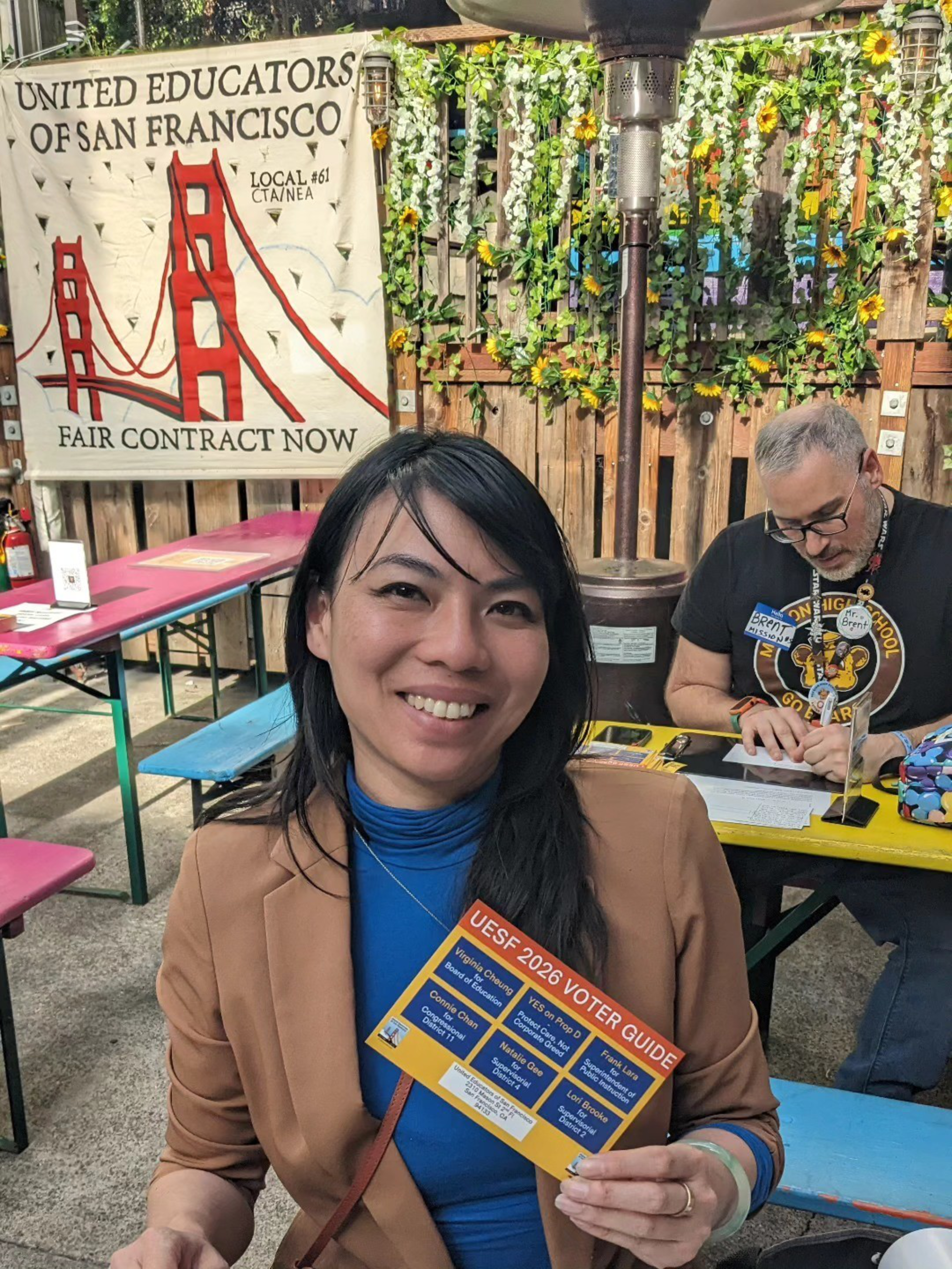 A woman smiles holding a UESF 2026 voter guide, sitting outside near tables with a “United Educators of San Francisco” banner and greenery behind her.