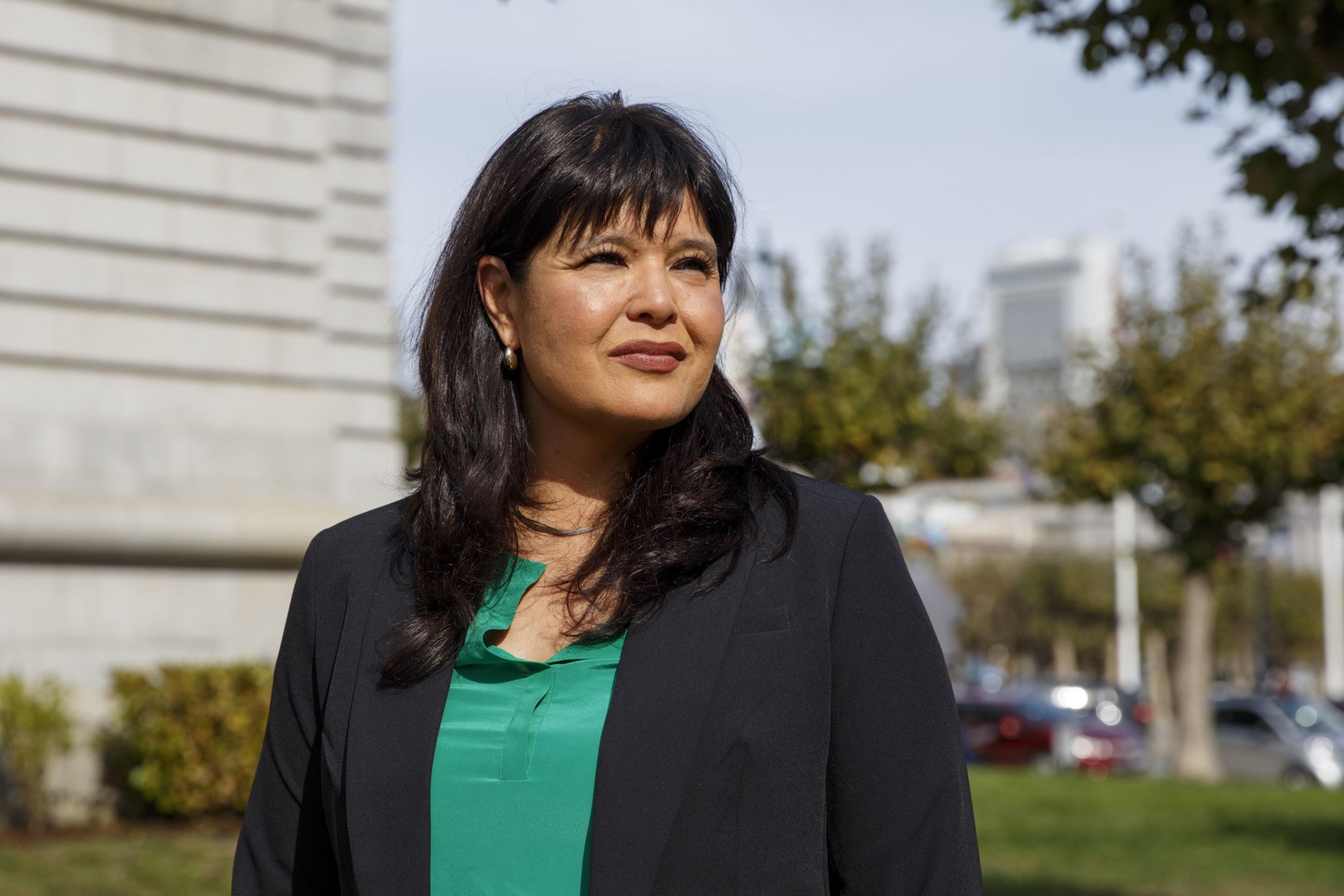 A woman outdoors, with dark hair, in a black jacket over a green blouse, slightly smiling. Trees and a building are in the background.