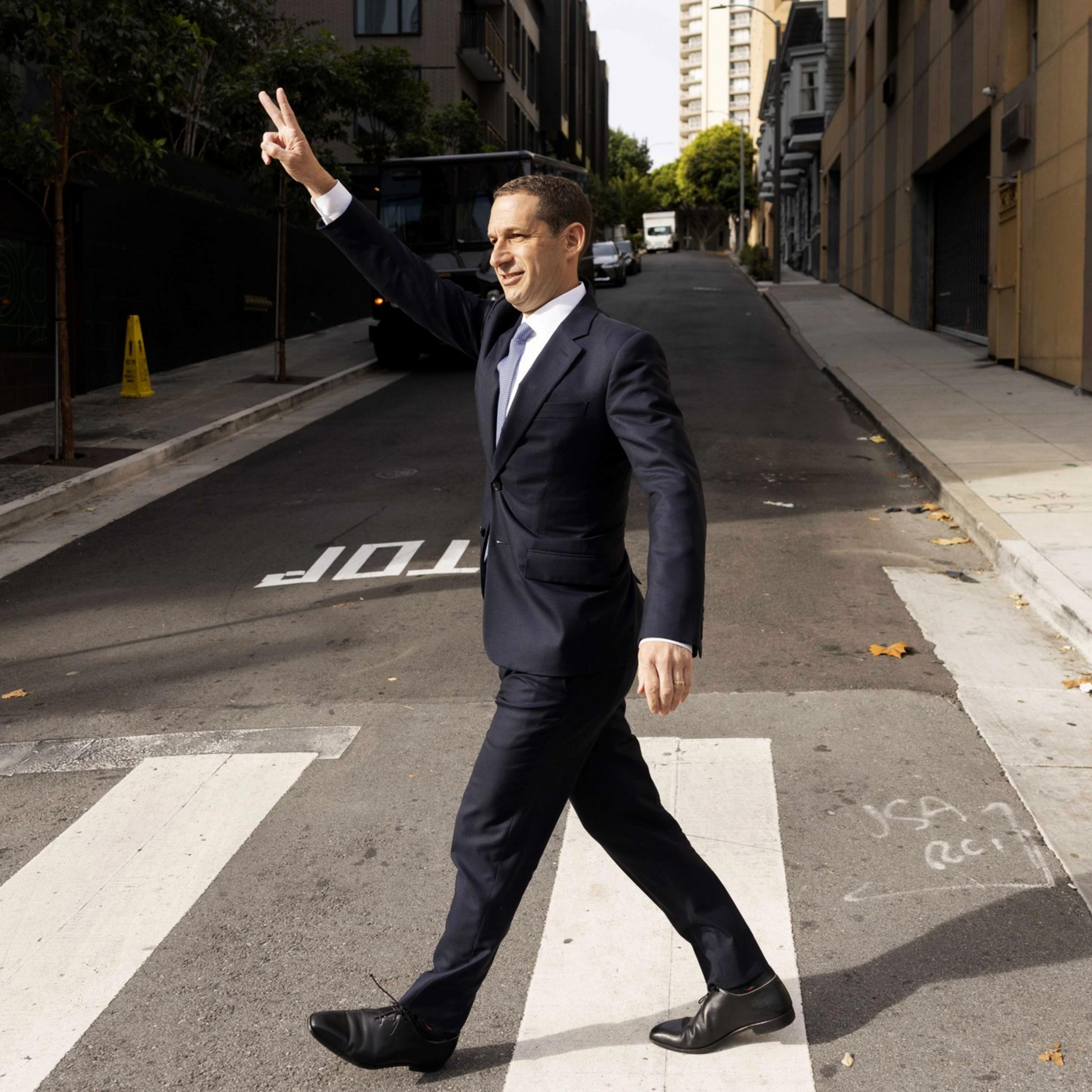 A man in a suit walks confidently across a crosswalk on a city street. He's raising his hand in a peace sign while smiling. The street is empty and lined with buildings.