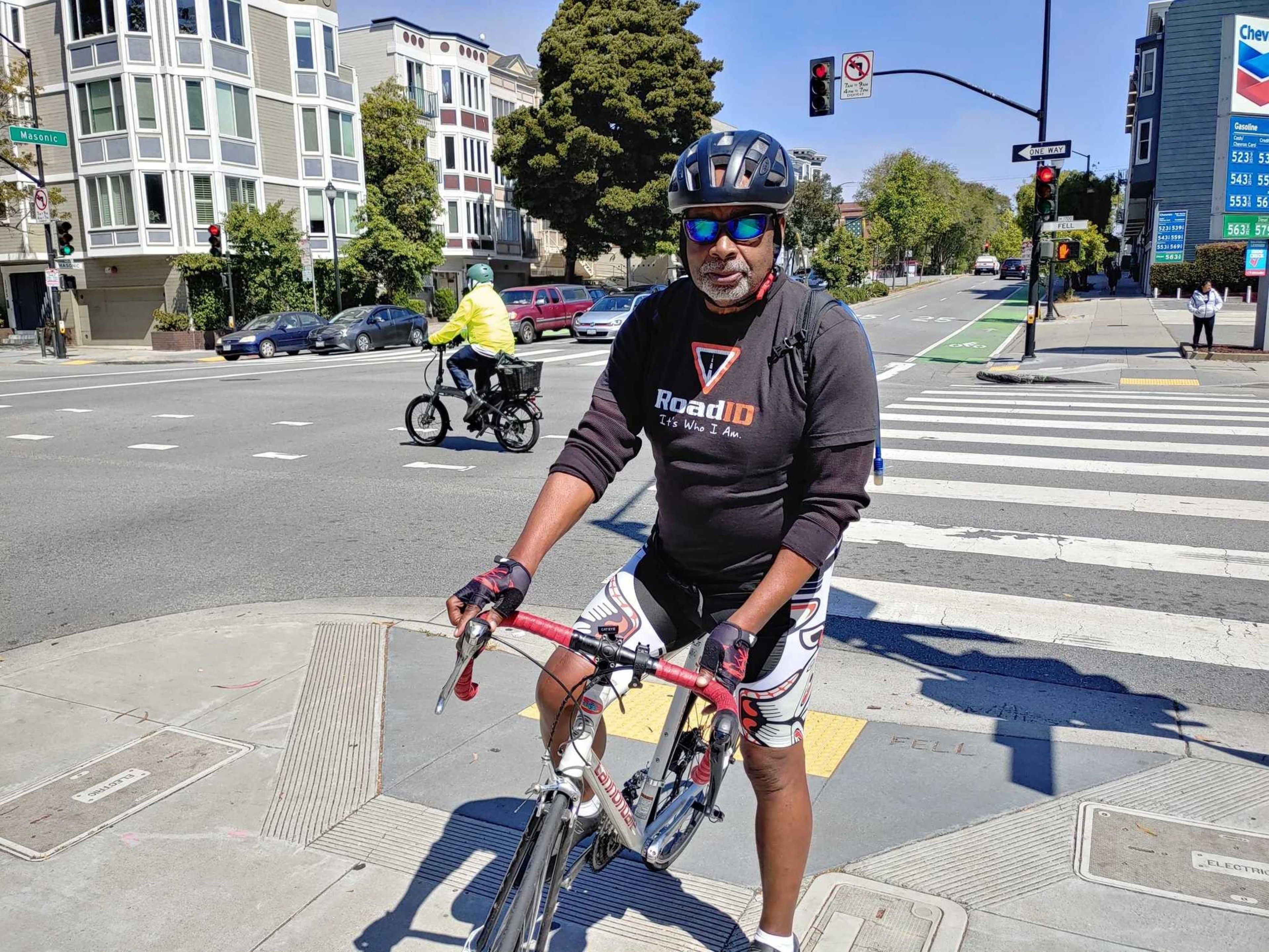 A man wearing a helmet and sunglasses is sitting on a road bike at a city intersection under a clear sky.