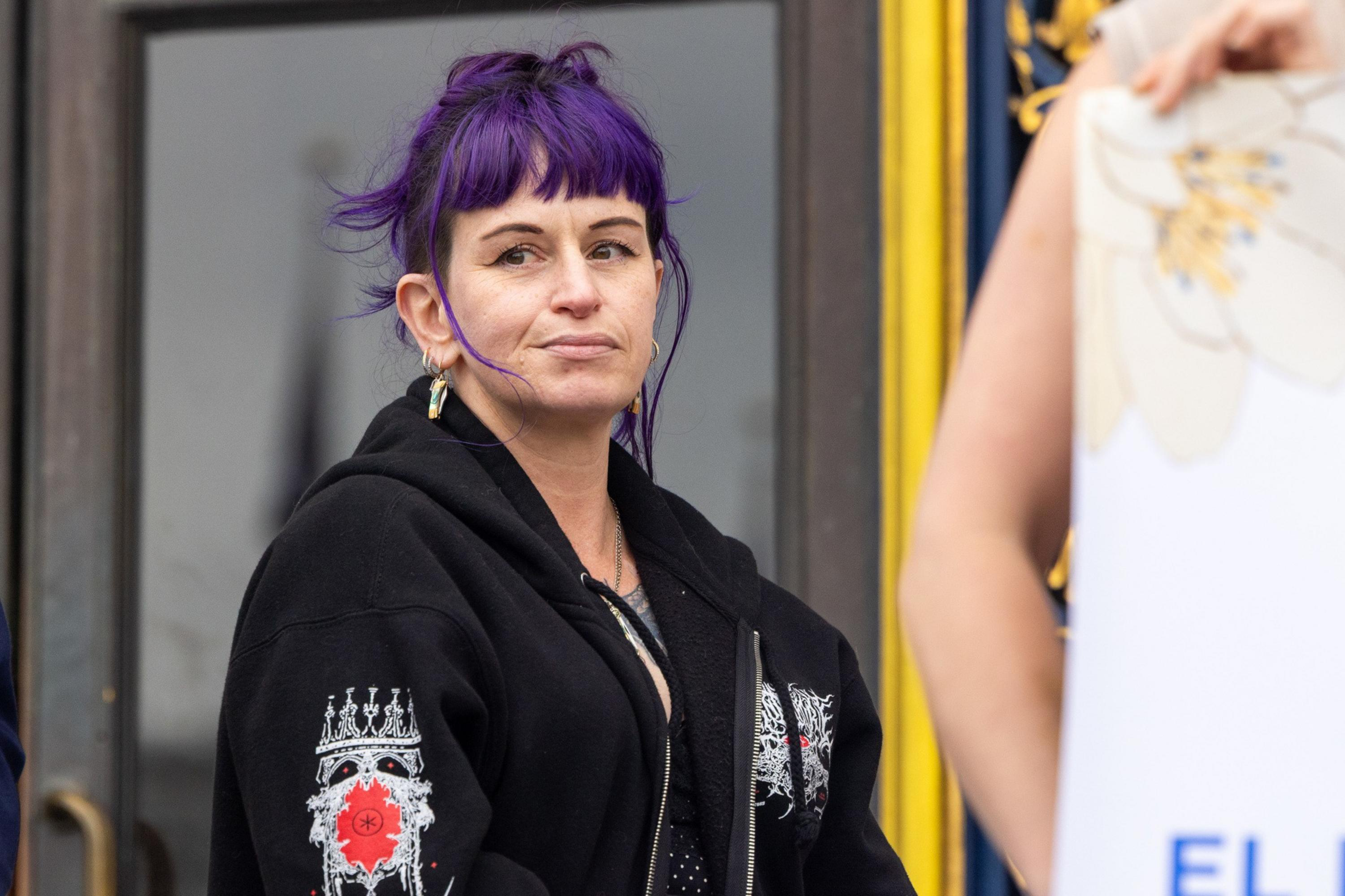 A woman with vibrant purple hair tied up, wearing a black hoodie with detailed white and red designs, looks to the side with a neutral expression.