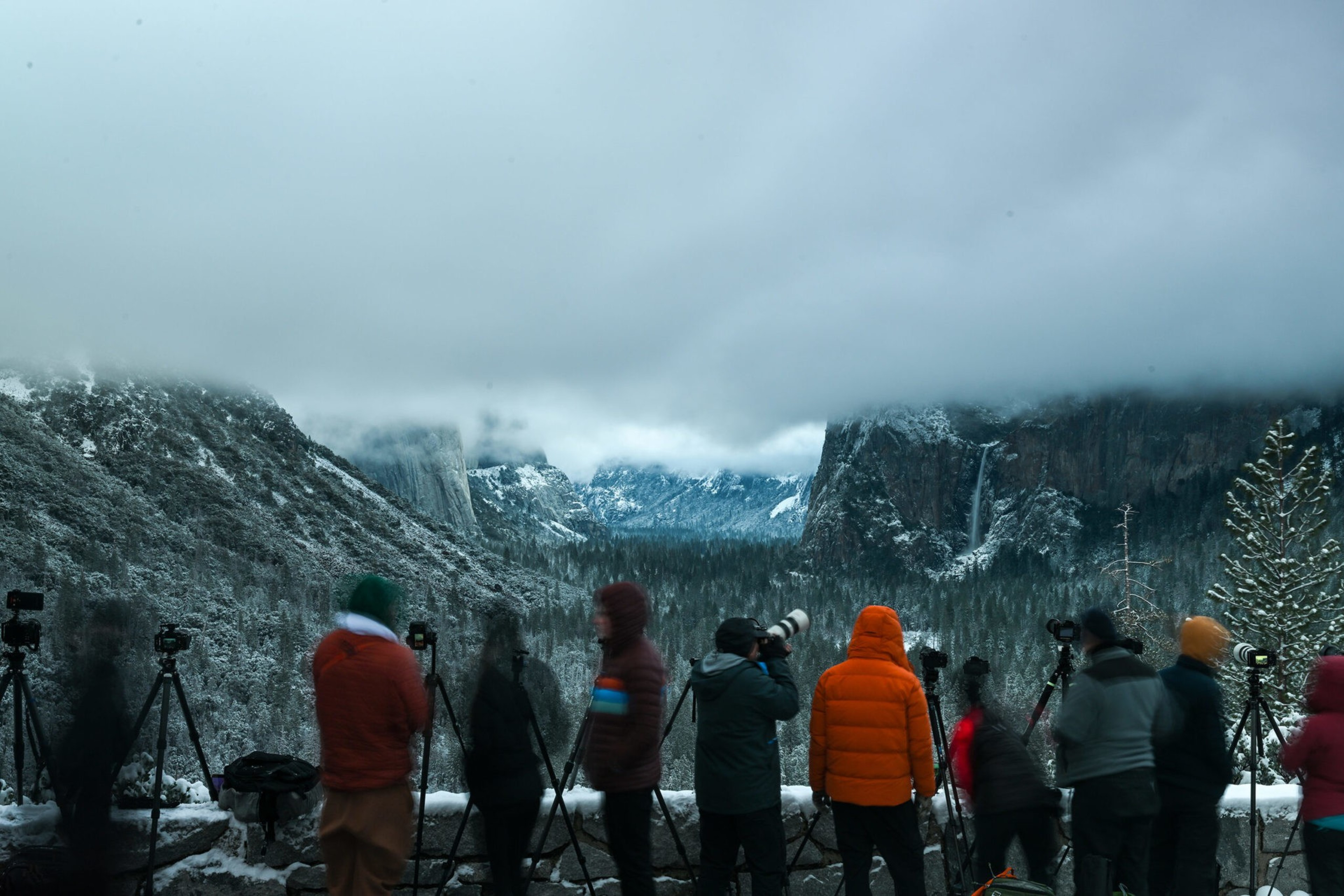 Several photographers in winter clothing stand on a snowy overlook, capturing a misty mountain valley with snow-covered cliffs and waterfalls under low clouds.