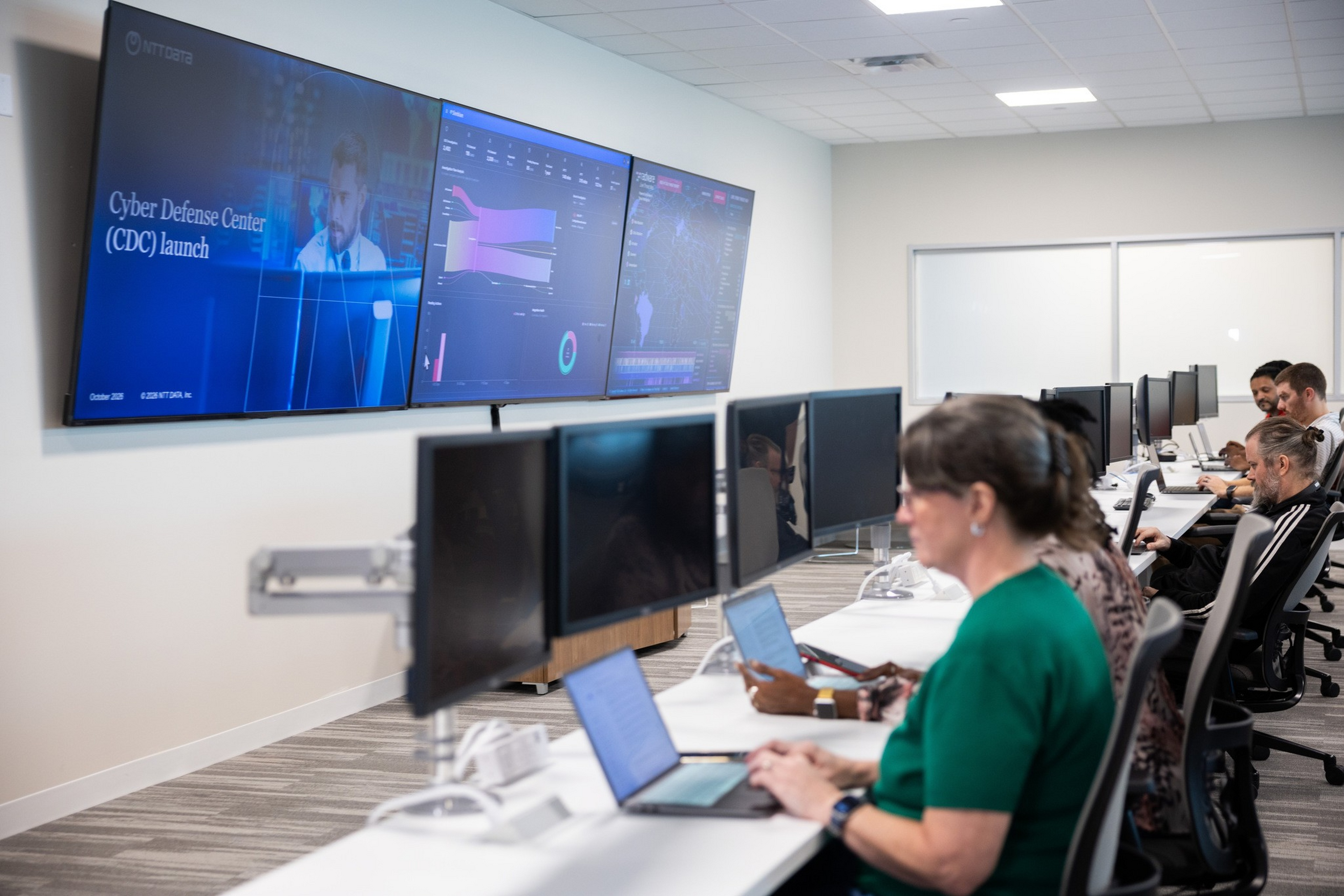 People work on laptops and desktops in a modern cybersecurity center with three large screens displaying data and a "Cyber Defense Center (CDC) launch" message.