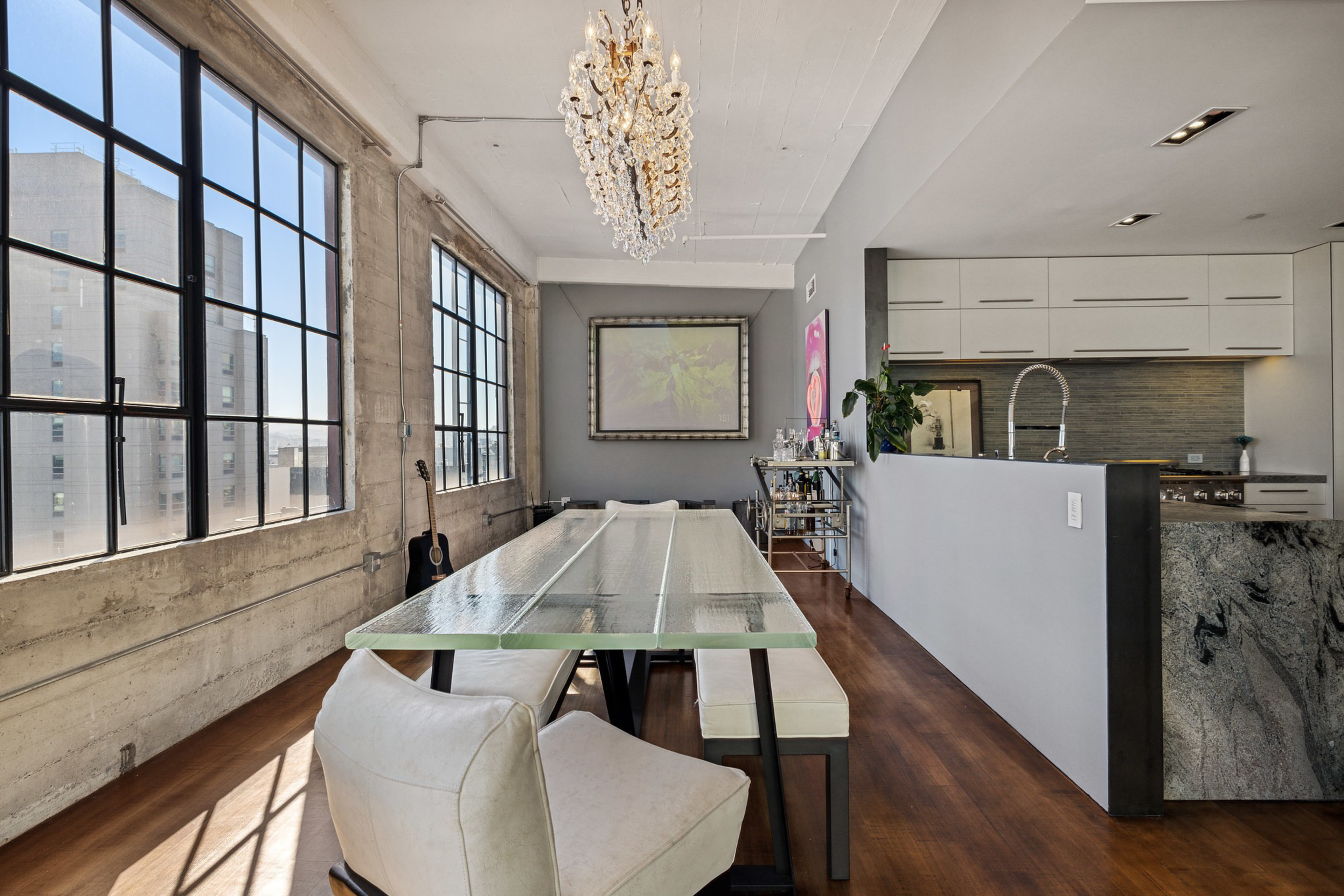 A modern dining area features a long glass table, white cushioned chairs and benches, large industrial windows, a chandelier, and an adjacent kitchen with white cabinets.