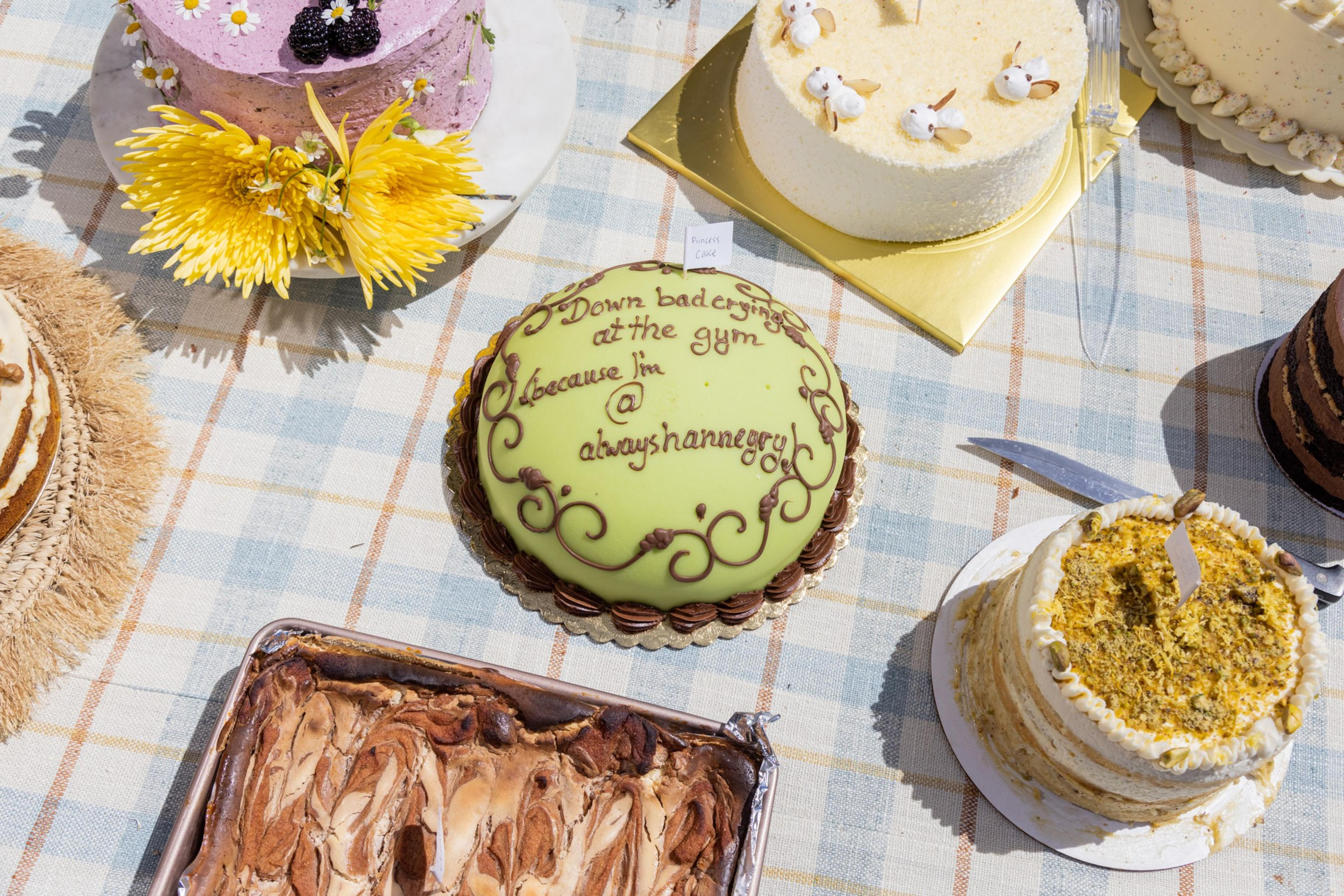 An assortment of cakes on a checkered cloth, one with a humorous gym-related message.