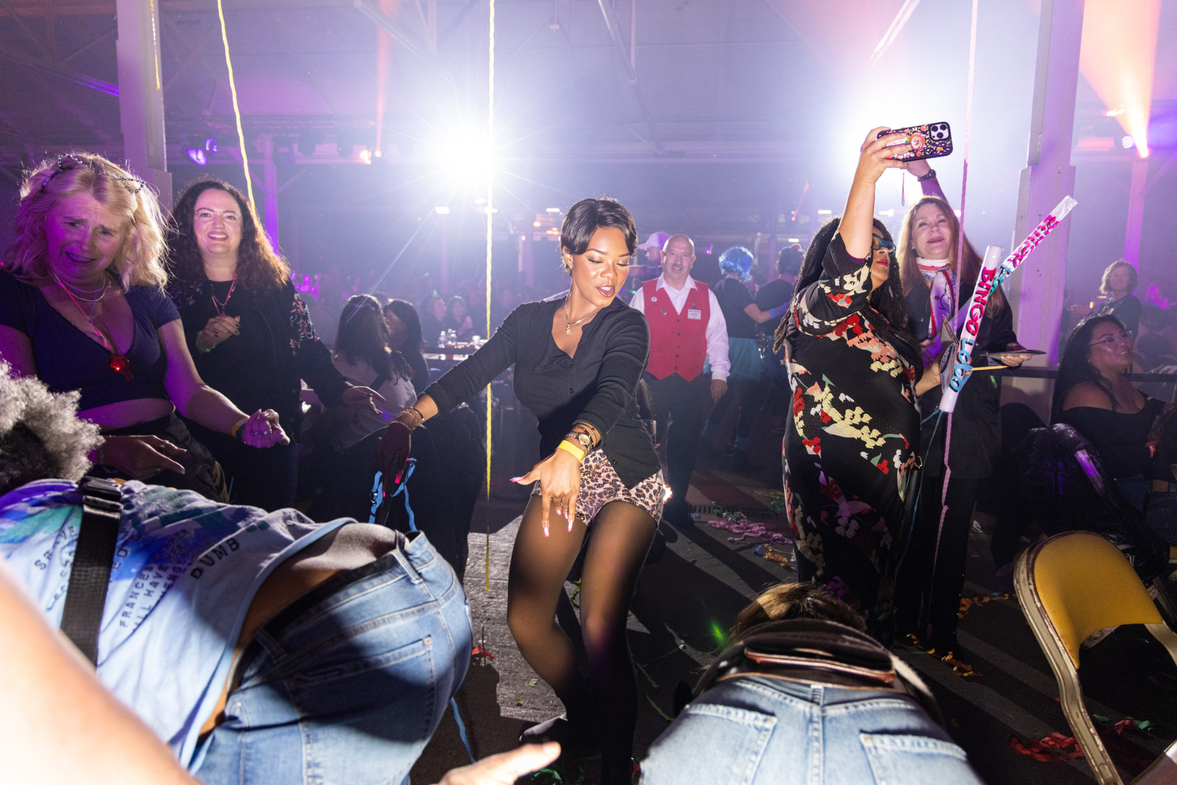 A lively party scene with people dancing, one woman taking a selfie, colorful lighting, and a festive atmosphere.