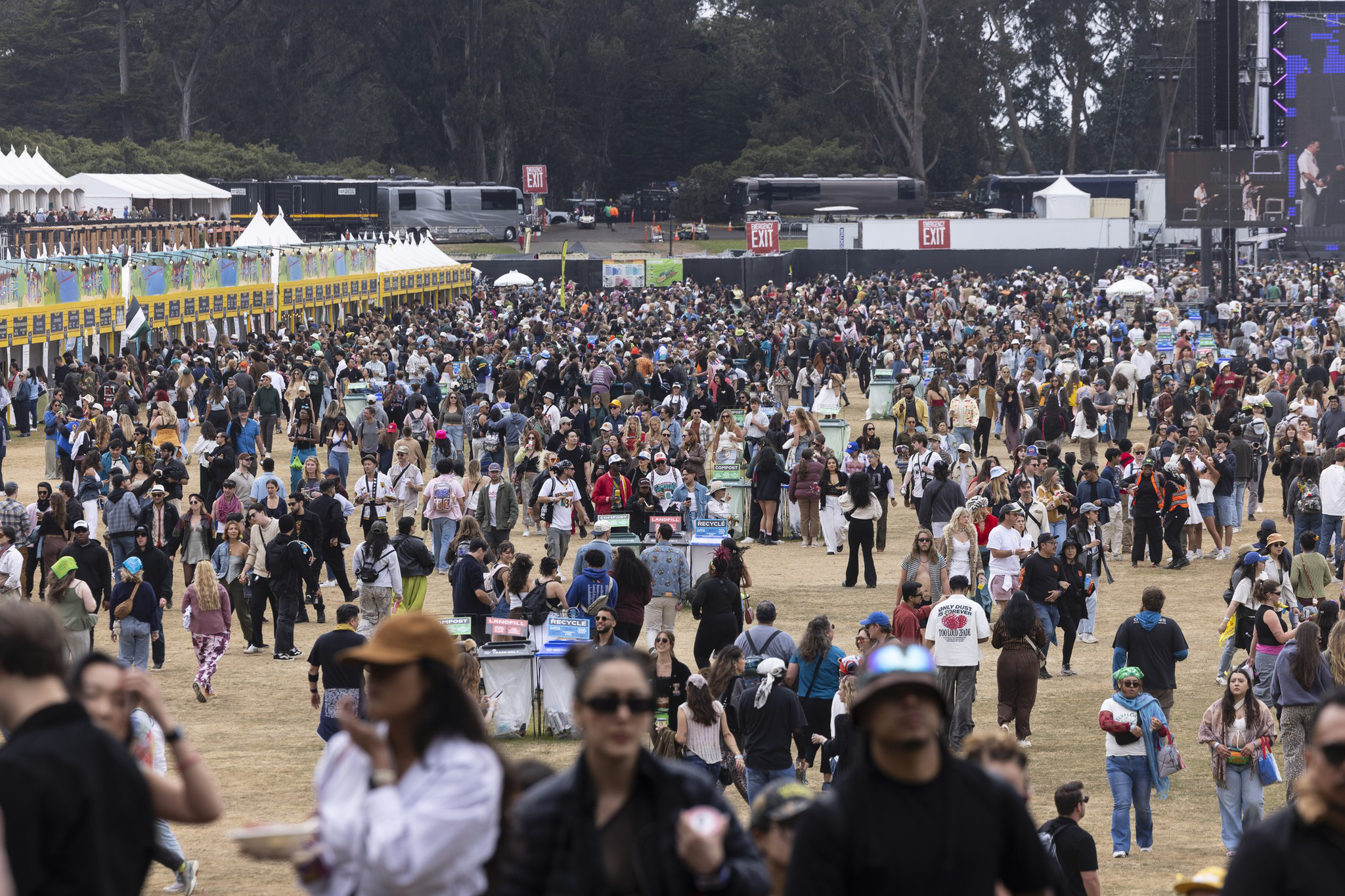 A large crowd of people gathers outdoors at a daytime event, with food stalls on the left and buses and exit signs in the background near tall trees.
