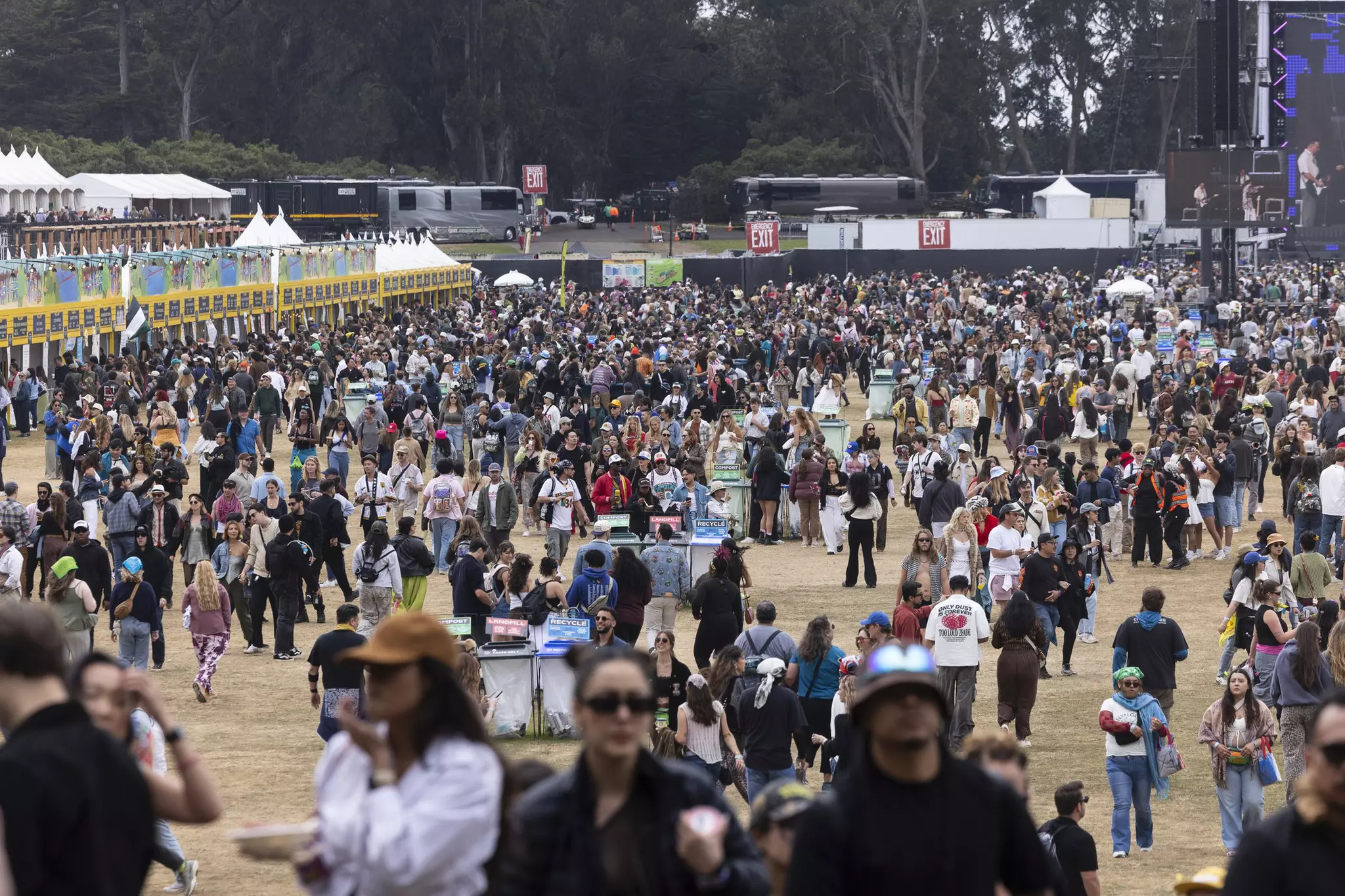 A large crowd of people gathers outdoors at a daytime event, with food stalls on the left and buses and exit signs in the background near tall trees.