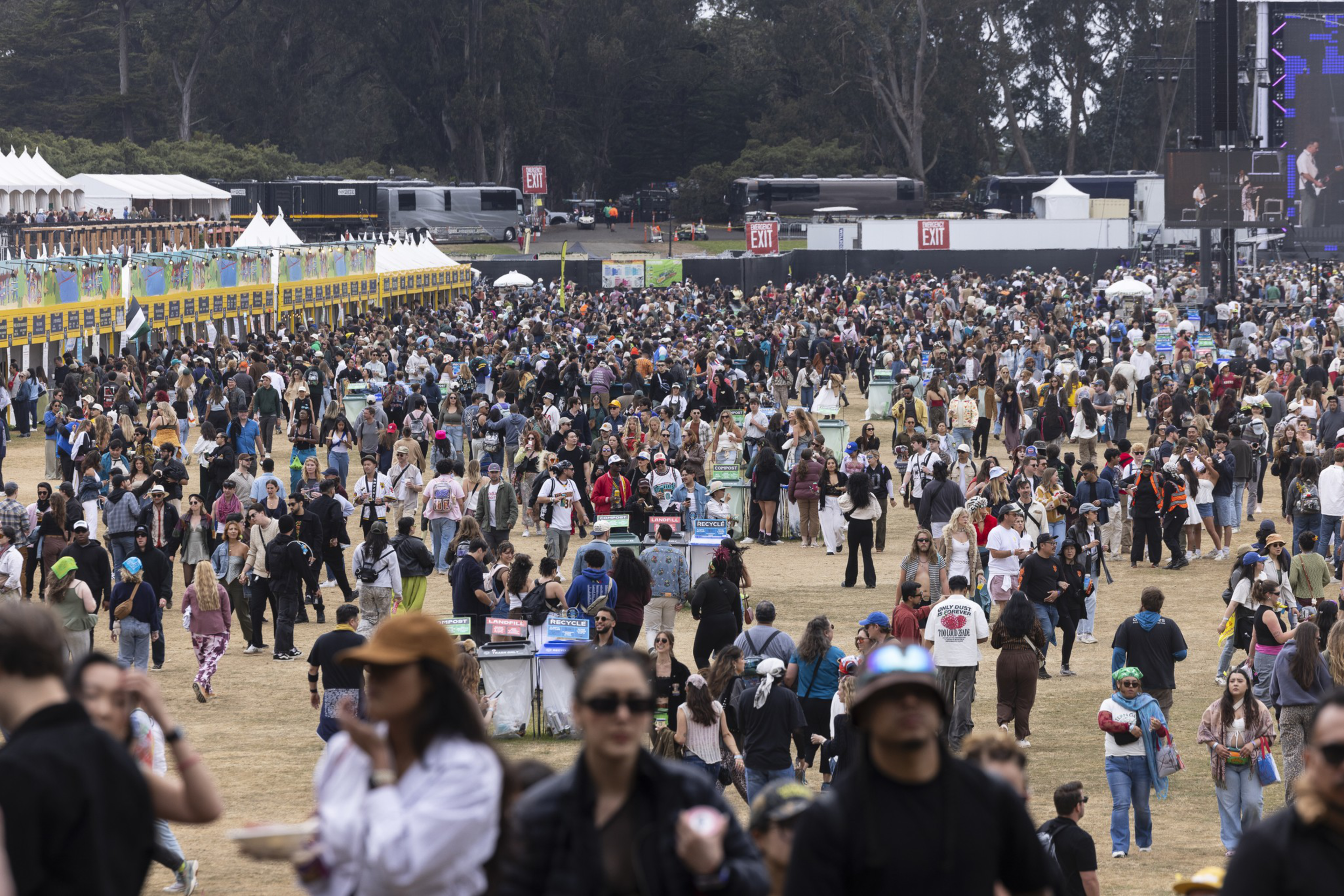 A large crowd of people gathers outdoors at a daytime event, with food stalls on the left and buses and exit signs in the background near tall trees.
