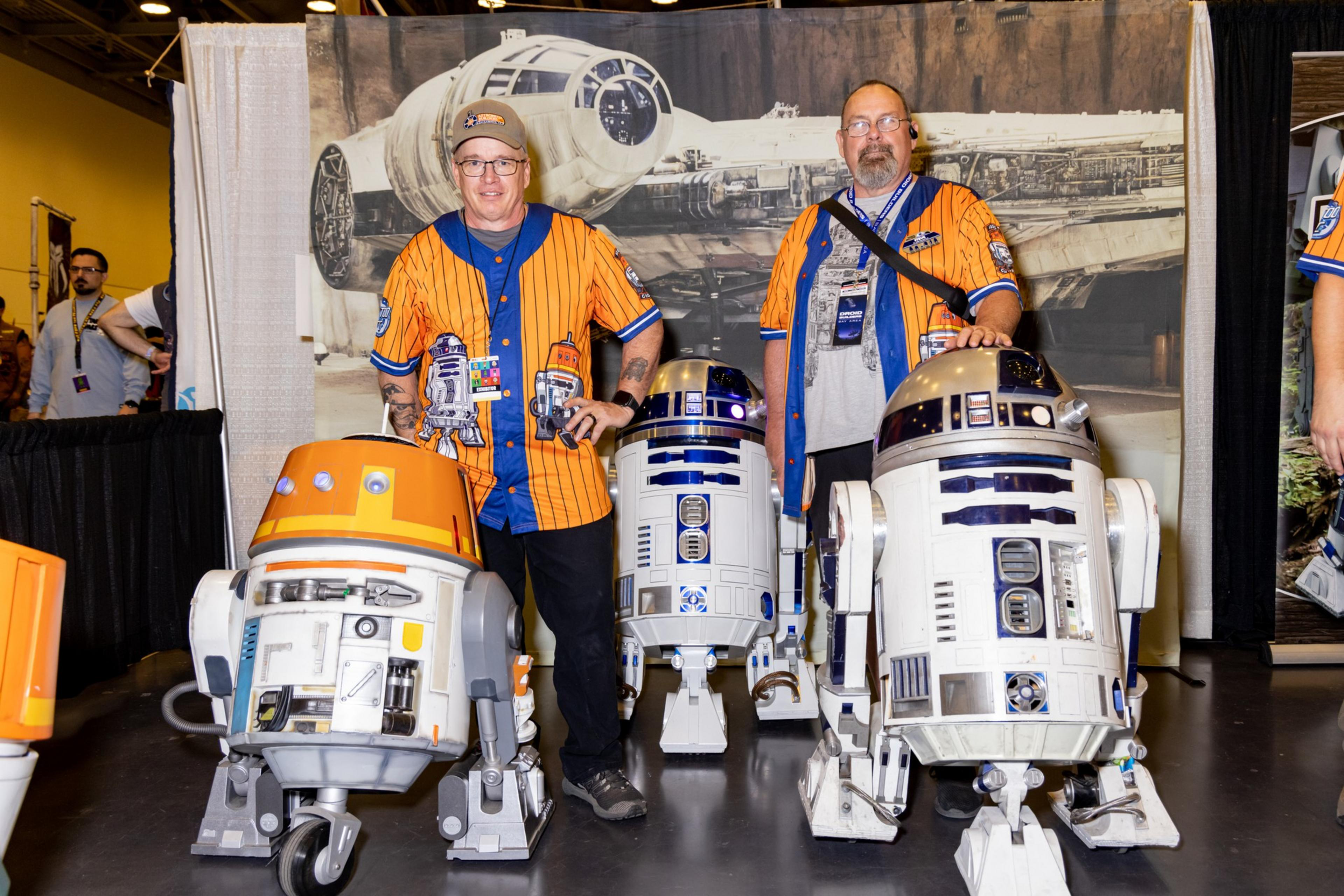 Two people stand smiling with custom robot replicas and props. They wear matching orange and blue uniforms with robot designs, in front of a sci-fi backdrop.
