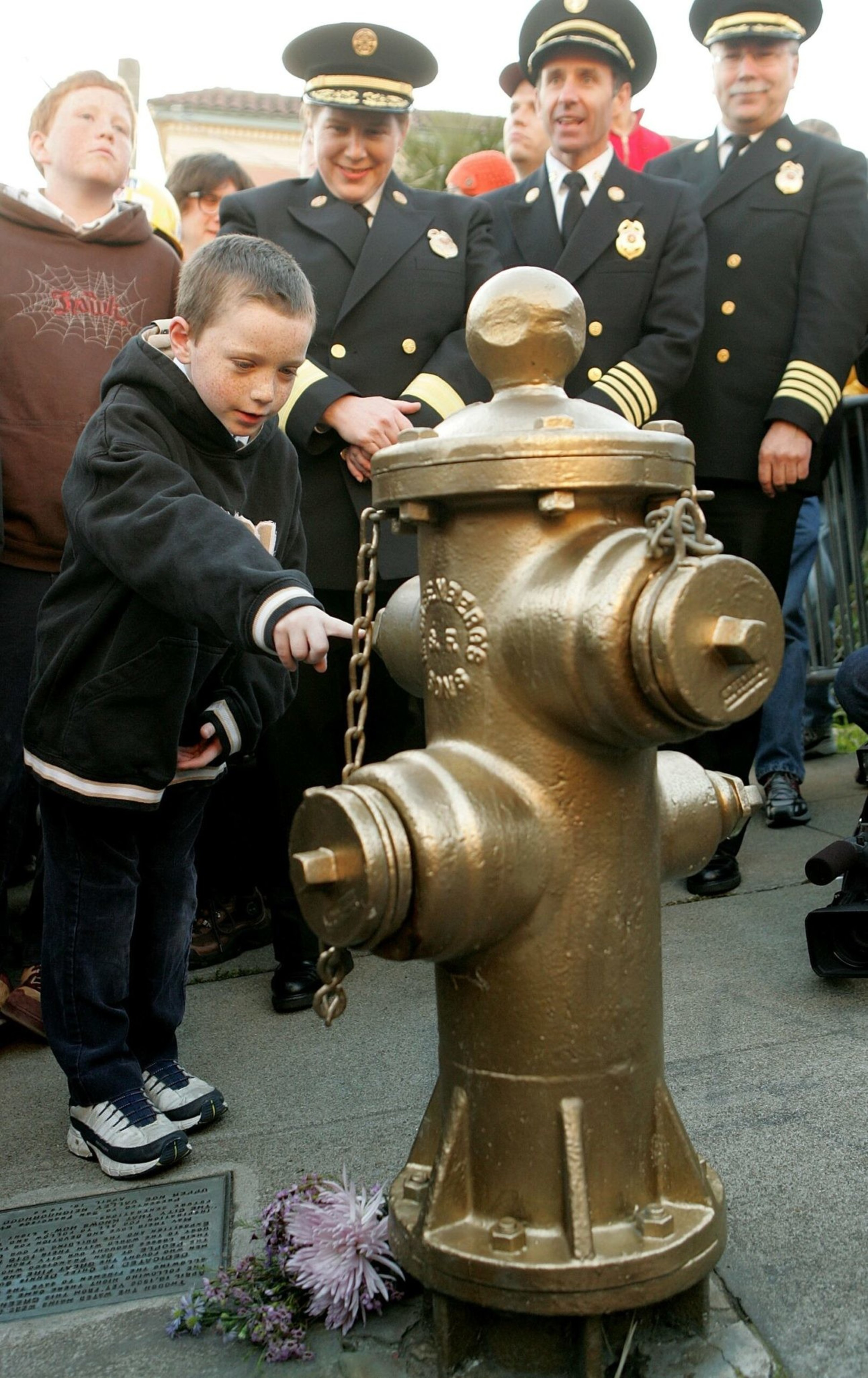 A young boy touches a gold-painted fire hydrant while firefighters in uniform and onlookers watch, with flowers placed at the hydrant’s base.