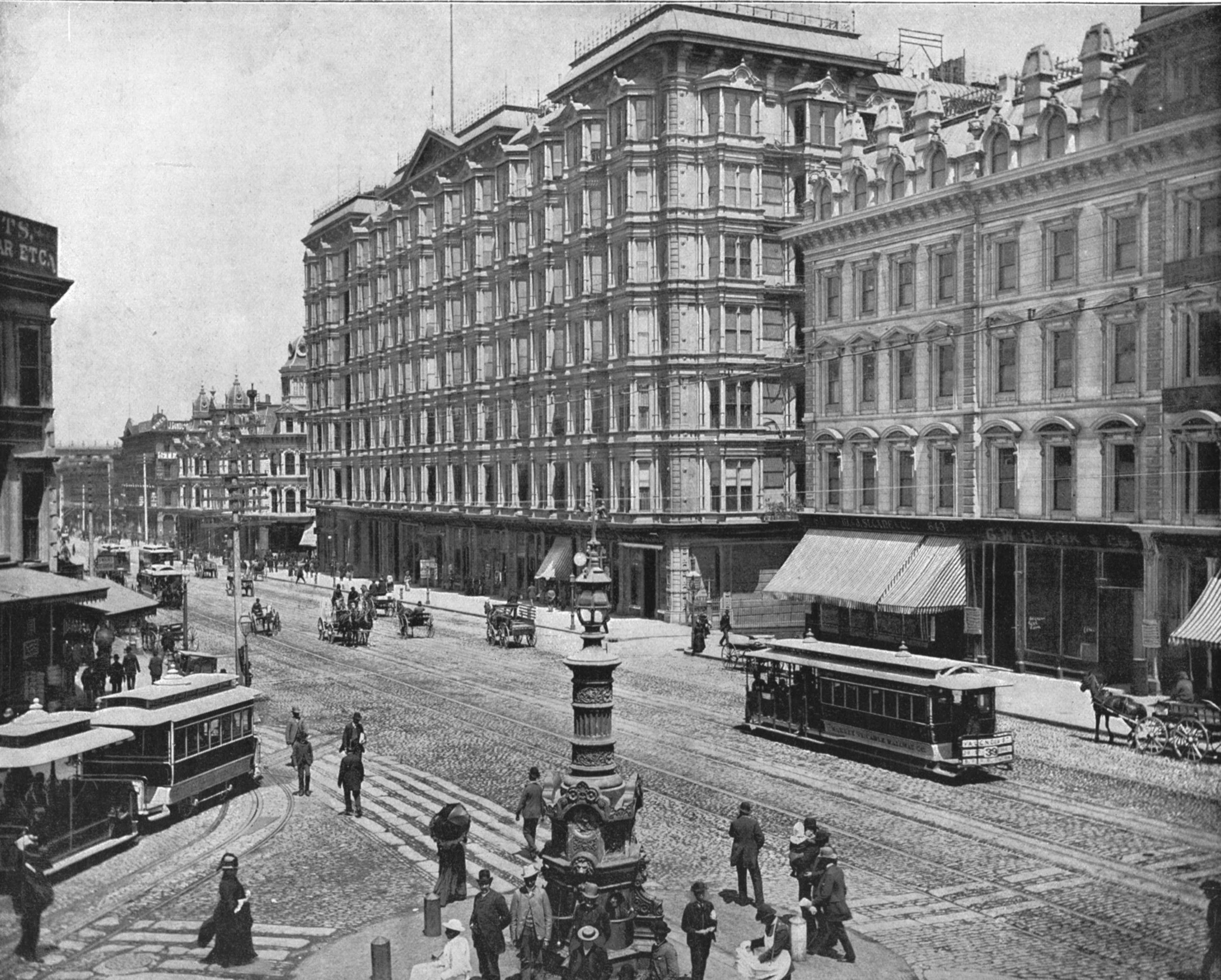 A busy city street with horse-drawn carriages, early streetcars, and pedestrians near tall, ornate buildings and a decorative street lamp.