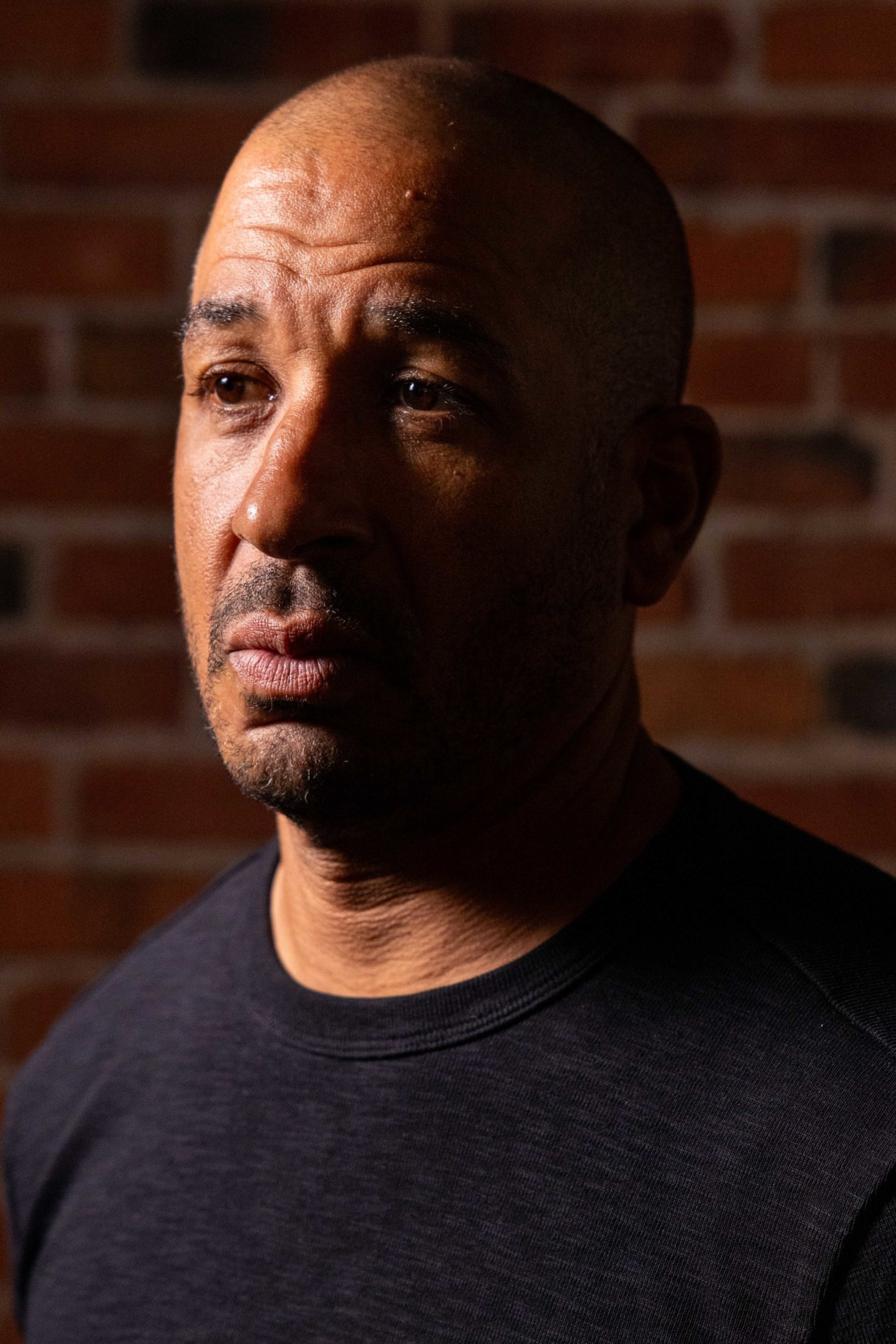 A bald man with a contemplative expression looks slightly to the side, lit by warm light against a blurred brick wall background.