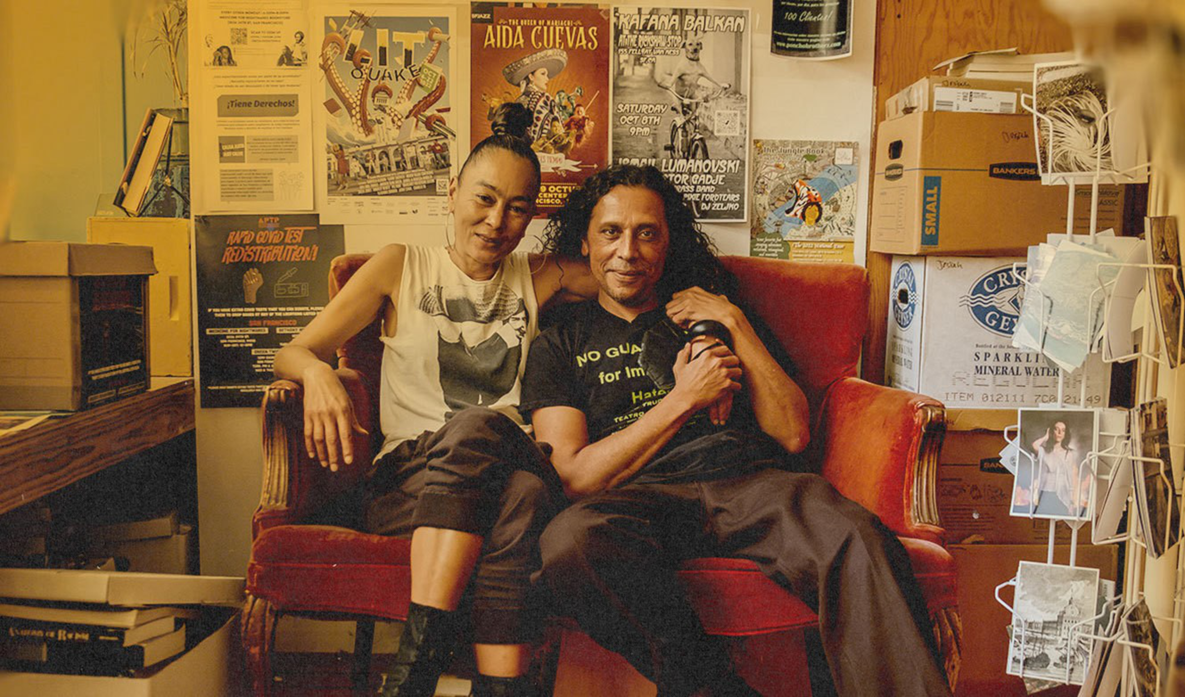 Two people sit closely on a red velvet chair in a cluttered room filled with posters, boxes, and a rack of photographs.